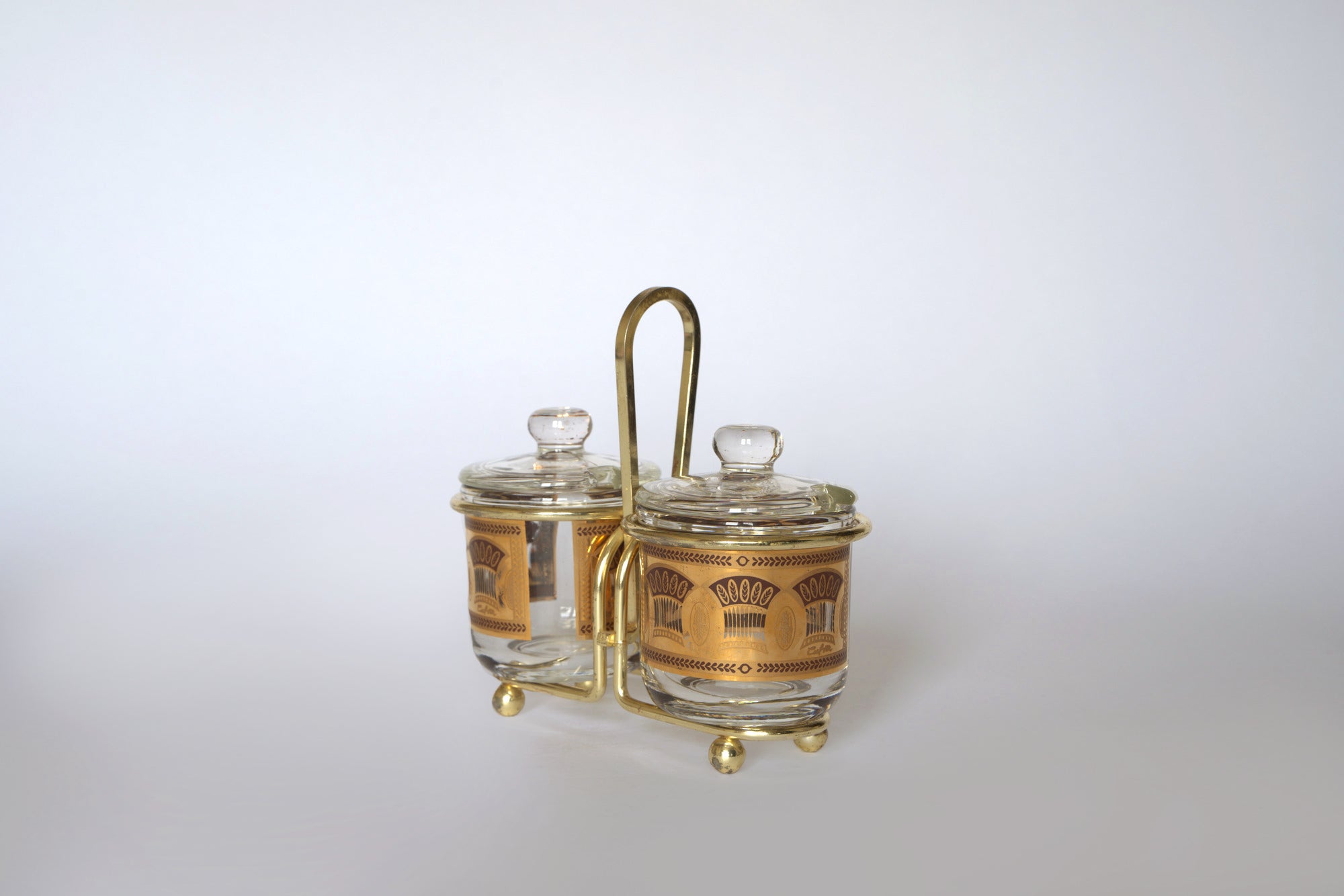The Vintage Gold Caddy Double Glass Jar Set by Euro Trash features two glass jars with gold patterns and clear lids in a gold metal holder with handle—a perfect vintage condiment set for mid-century decor.