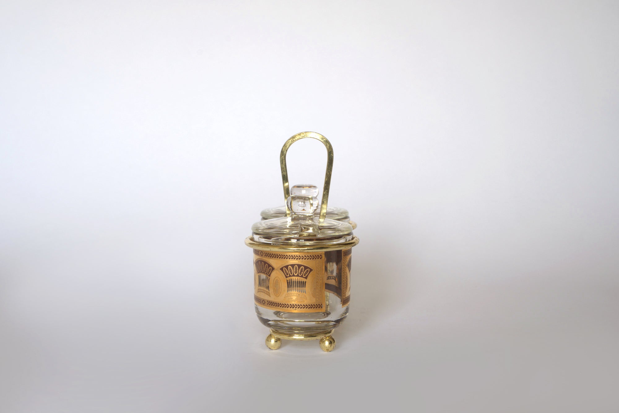 The Euro Trash Vintage Gold Caddy Double Glass Jar Set features clear glass jars with decorative gold accents and handles, perfect for vintage condiment sets or adding mid-century charm to your kitchen decor.