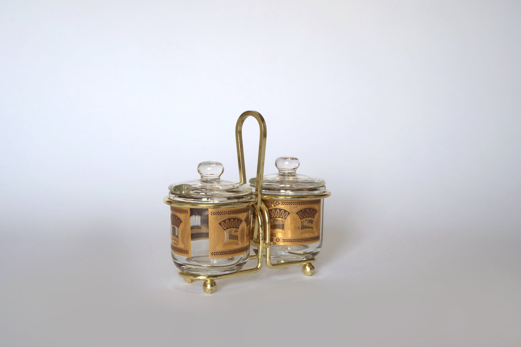 The Vintage Gold Caddy Double Glass Jar Set by Euro Trash features two small glass jars with gold and brown designs in a metal holder—perfect for adding retro charm to your mid-century kitchen.