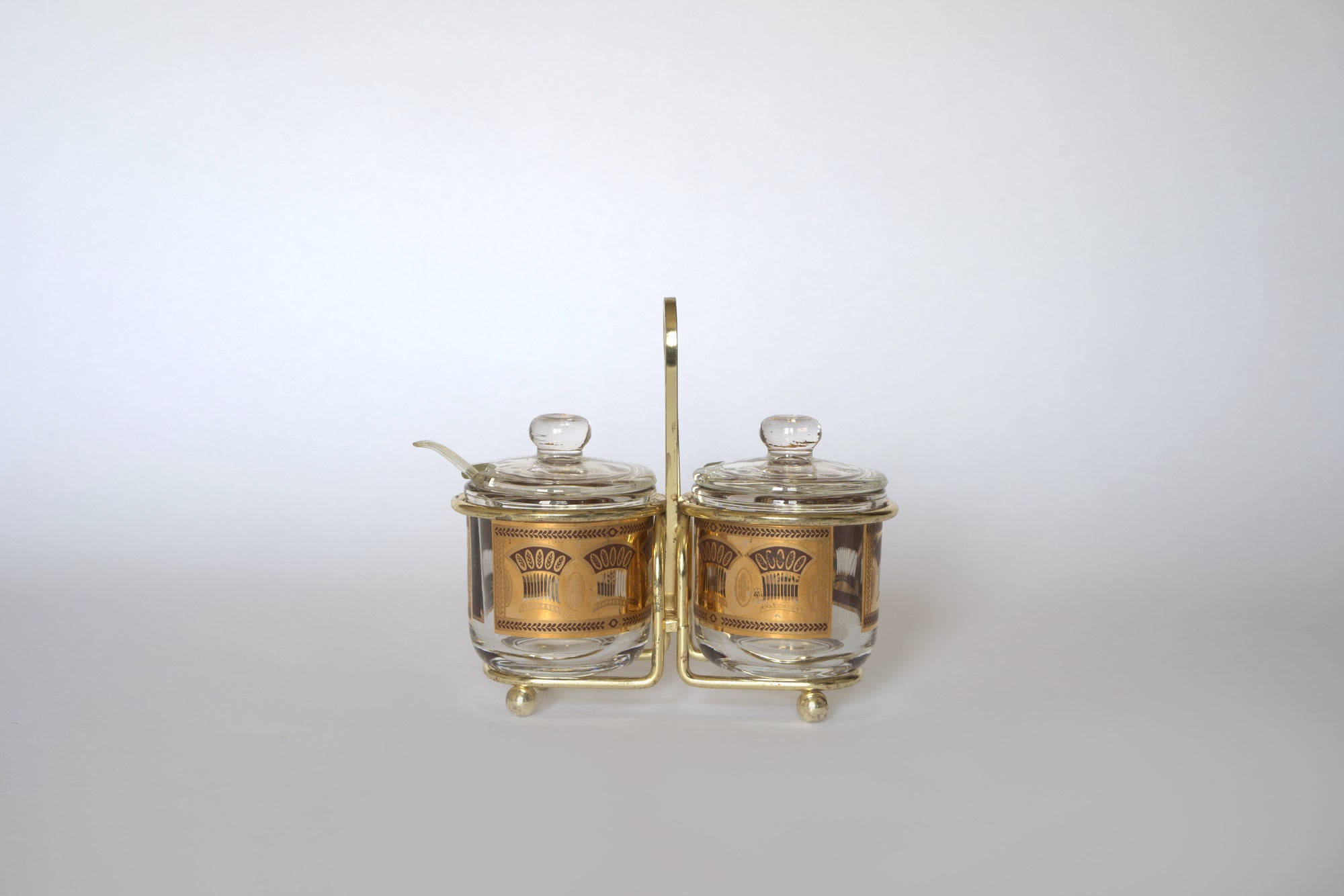 The Euro Trash Vintage Gold Caddy Double Glass Jar Set features two patterned jars with lids and gold trim, set in a handled gold holder. One jar includes a small spoon. Perfect for retro kitchen decor; background is plain and light-colored.