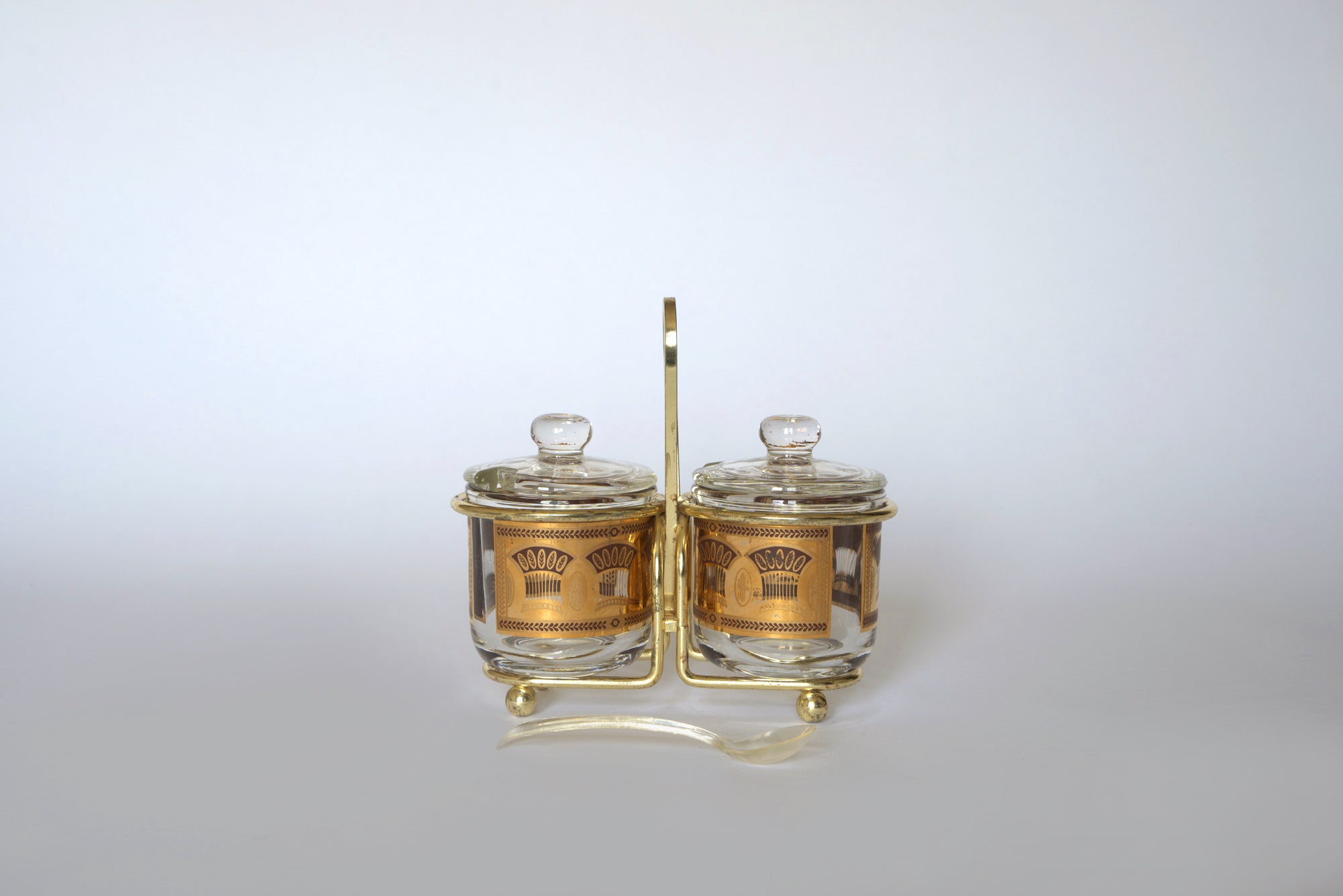 The Euro Trash Vintage Gold Caddy Double Glass Jar Set includes two glass jars with gold accents and lids in a gold-tone metal stand with handle, plus a clear plastic spoon—ideal for adding mid-century style to your kitchen decor.