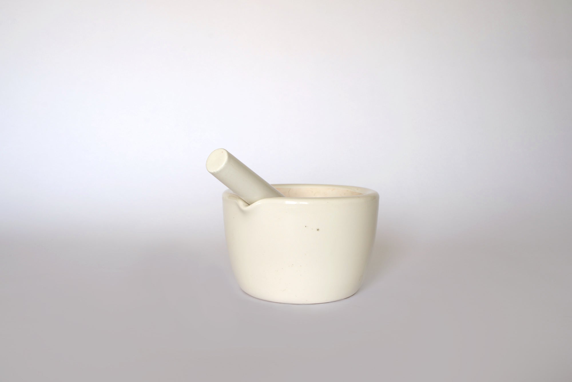 The Euro Trash Vintage White Ceramic Mortar and Pestle Set sits on a plain, light surface with a neutral background. The pestle rests inside the mortar, making this set an ideal decorative accent for your kitchen.