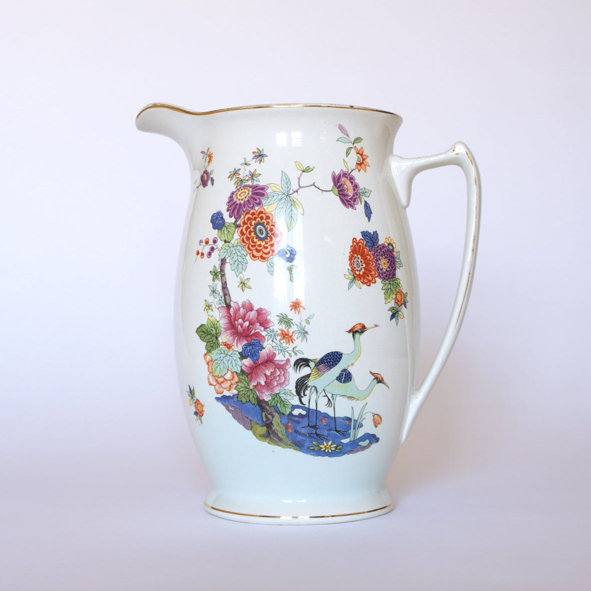 The Vintage Floral Porcelain Pitcher by Euro Trash features a curved handle, vibrant floral designs, and elegant crane motifs by the water on a plain light background.