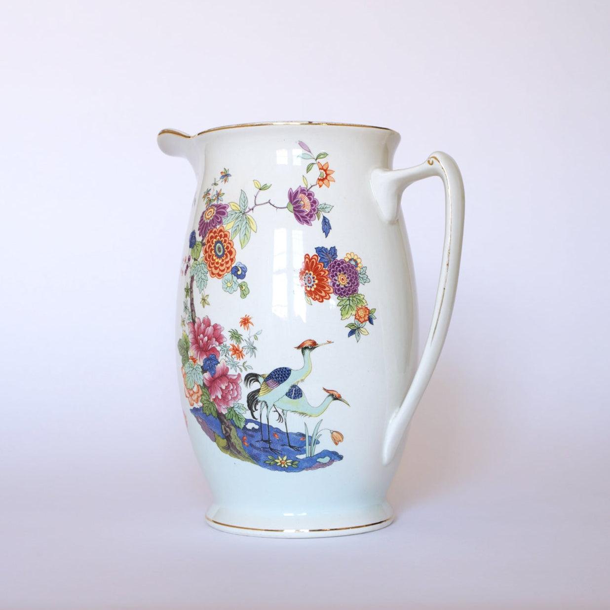 The Vintage Floral Porcelain Pitcher by Euro Trash features a curved handle, vibrant floral patterns, and elegant crane motifs near water on a plain light background.