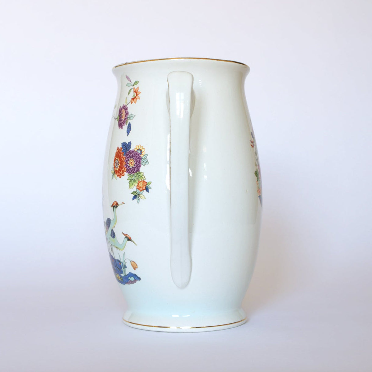The Vintage Floral Porcelain Pitcher by Euro Trash features a handle, colorful floral patterns, and delicate crane motifs on the side, all set against a plain white background.