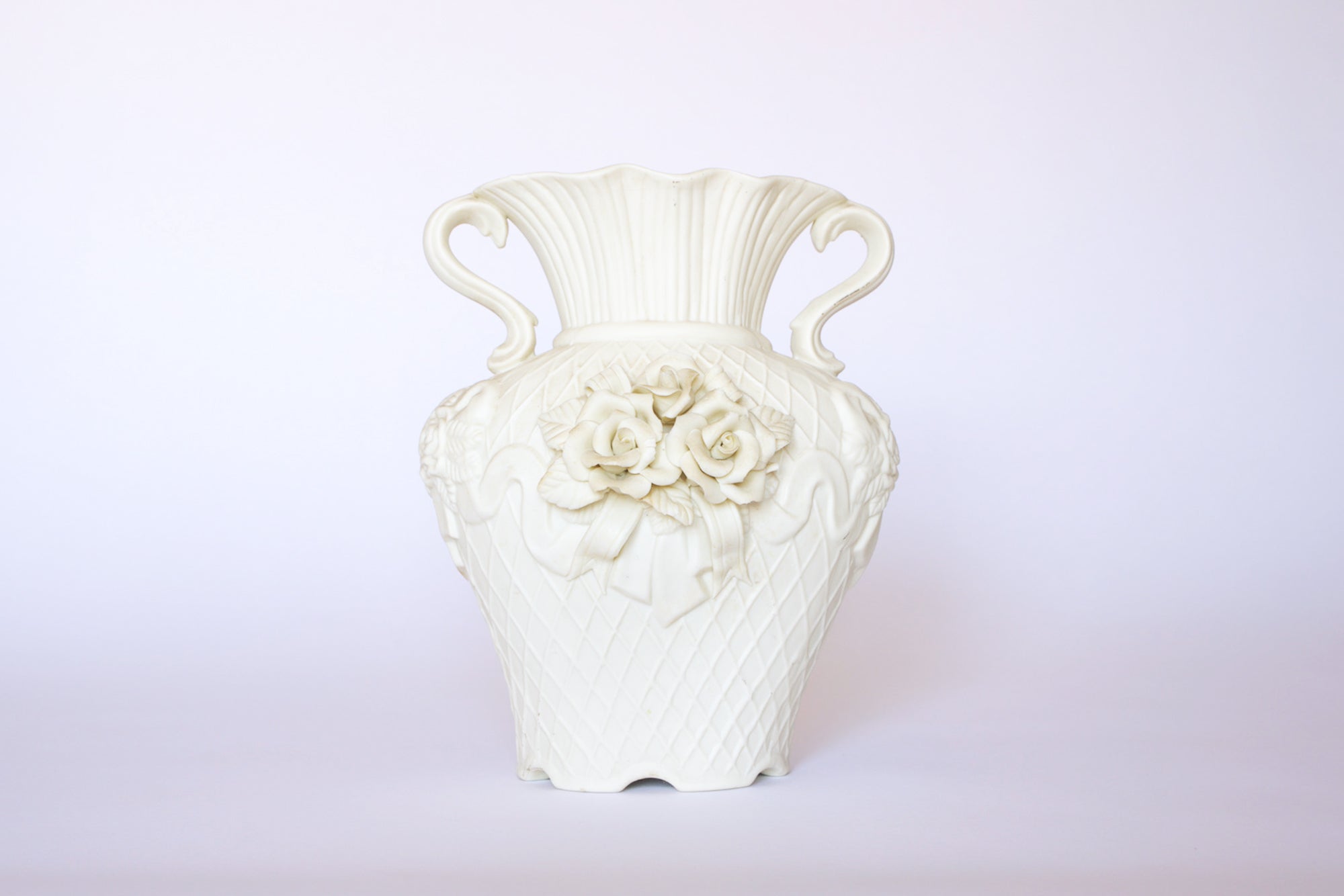 The Euro Trash Annie Vintage White Ceramic Relief Floral Vase features a quilted texture, decorative handles, and a sculpted bouquet of roses on the front, displayed against a plain light background.