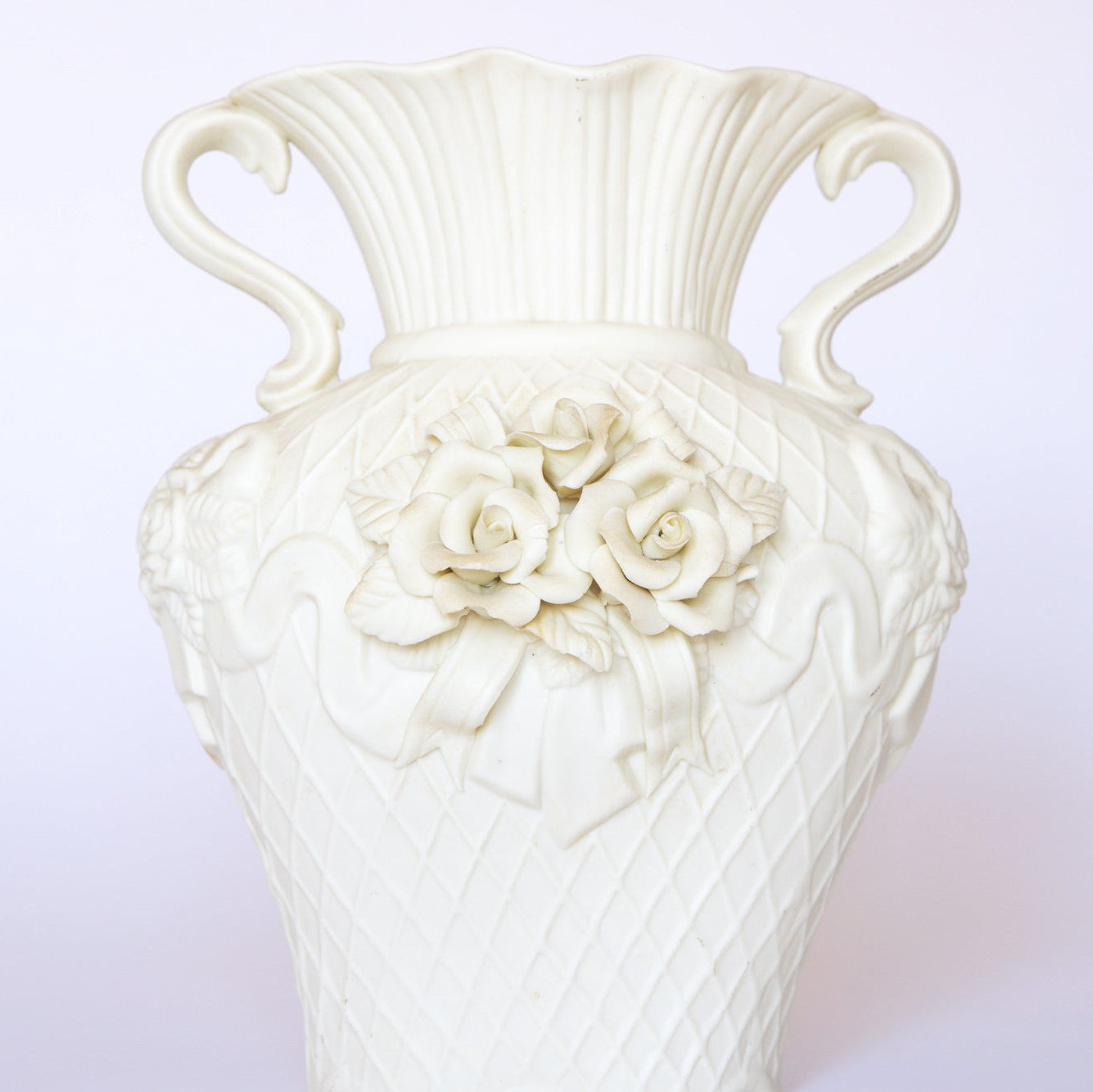 The Euro Trash Annie Vintage White Ceramic Relief Floral Vase features dual handles, embossed roses, ribbon accents, a diamond-patterned texture, and a fluted top—perfect for classic ornamental decor.