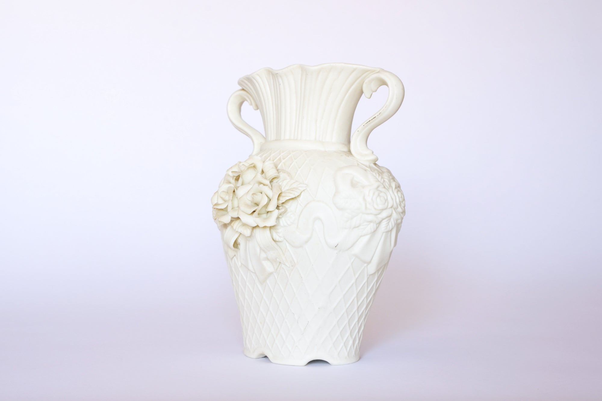 The Euro Trash Annie Vintage White Ceramic Relief Floral Vase features two handles, a scalloped rim, and detailed rose and leaf ornaments on the front, displayed against a plain white background.