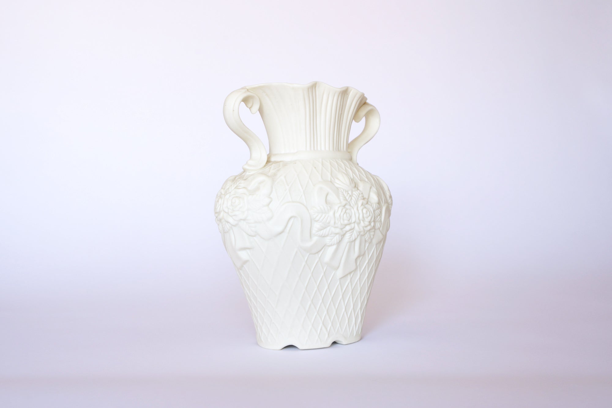 The Euro Trash Annie Vintage White Ceramic Relief Floral Vase features ornate floral detailing, a scalloped rim, and two decorative handles, shown against a plain light background.