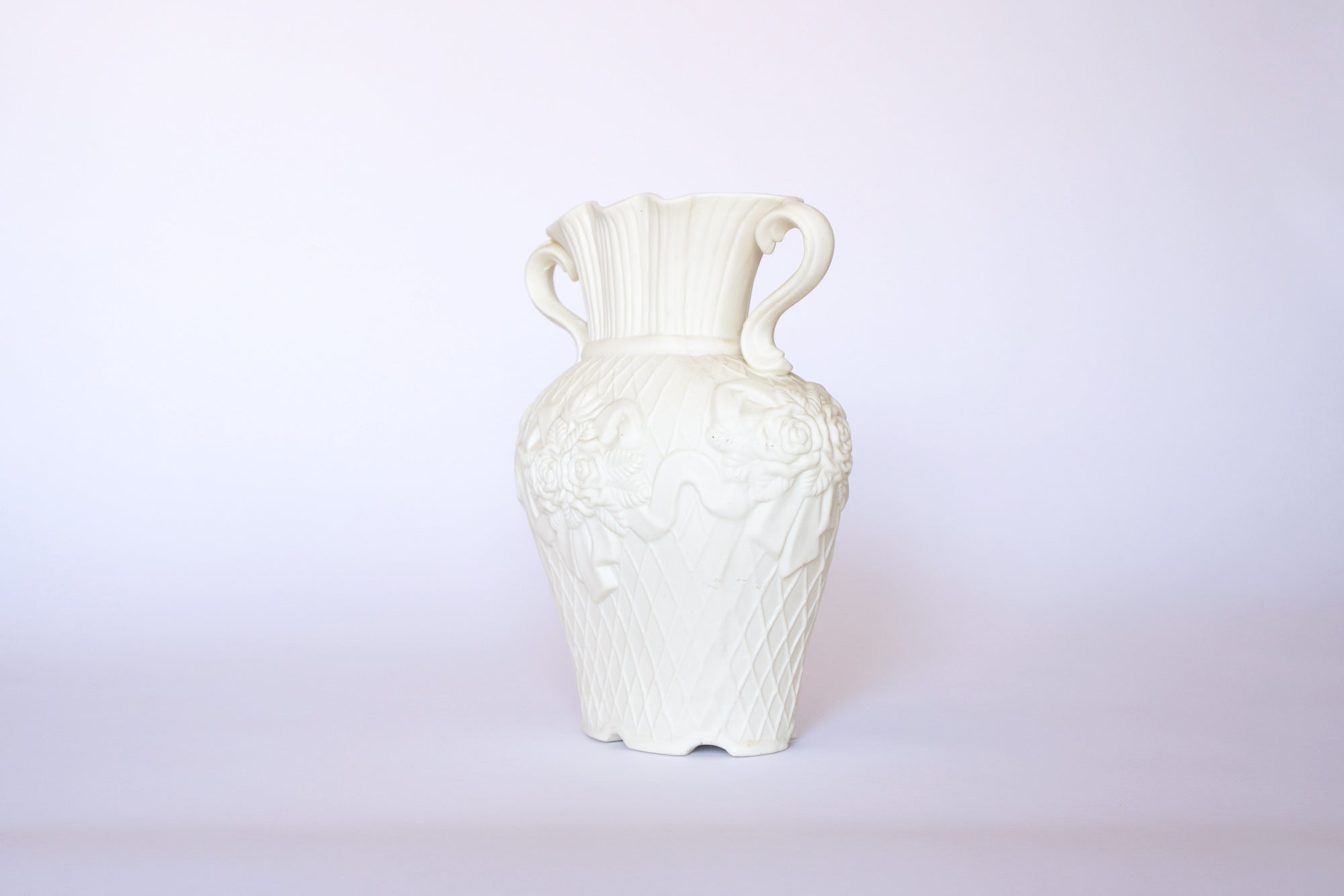 The Vintage White Ceramic Relief Floral Vase by Euro Trash Annie showcases ornate handles, intricate floral detailing, and lattice patterns, making it a striking ornamental decor piece against a light background.