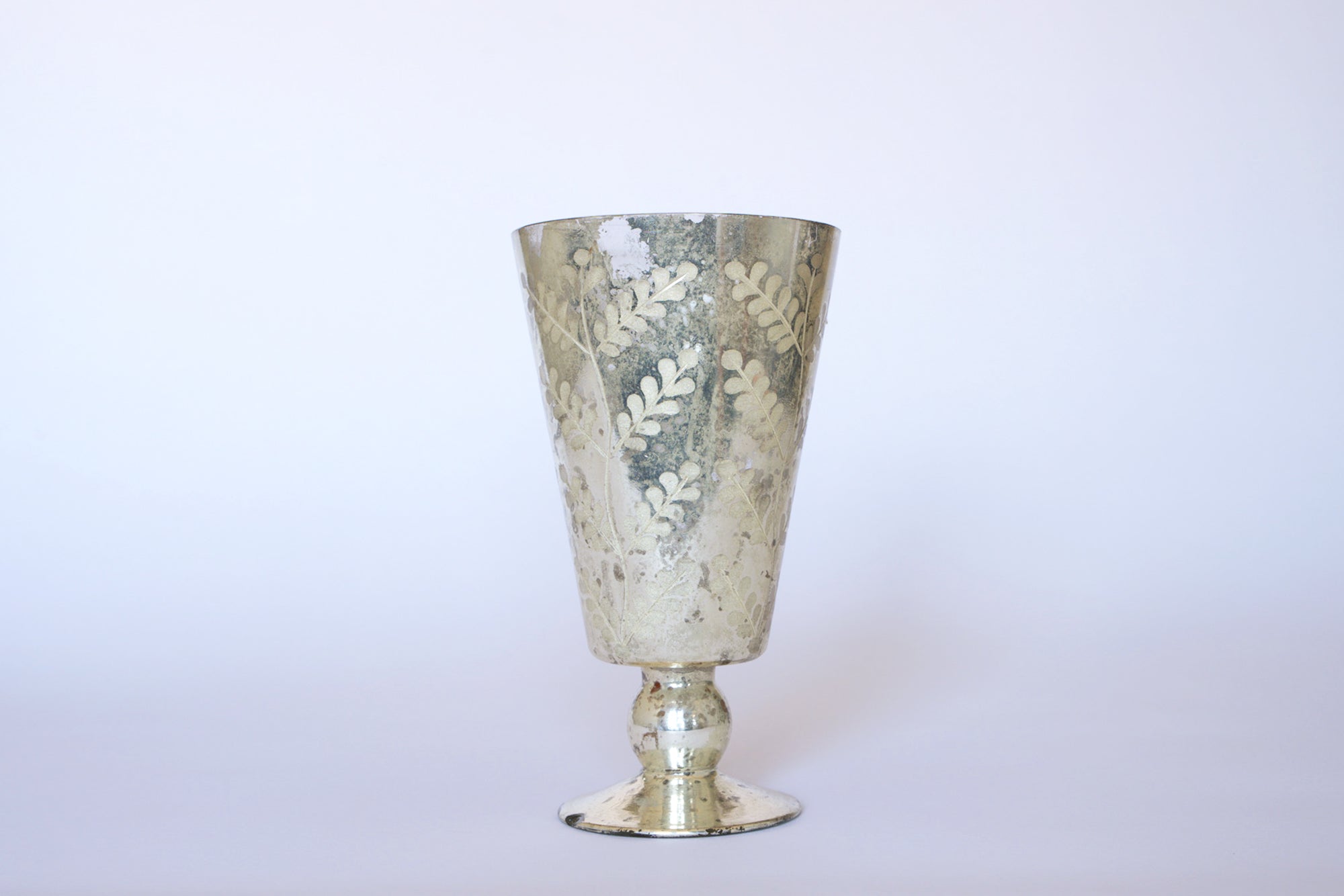 The Euro Trash Vintage Mercury Glass Vase features a metallic finish, flared rim, and etched leaf details on a simple pedestal base, beautifully capturing the charm of antique silver decor.
