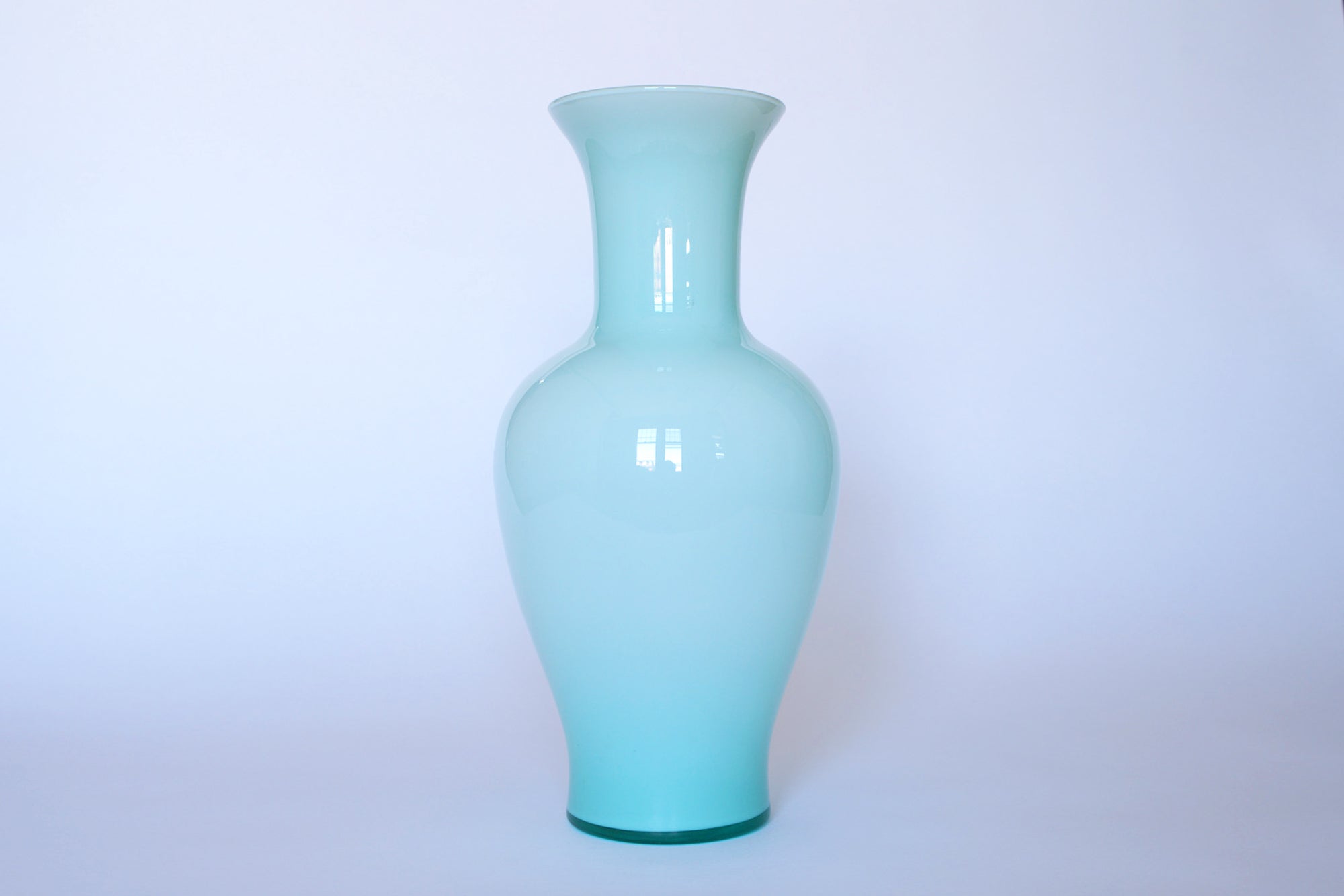 The Euro Trash Vintage Pale Blue Glass Vase features a tall, glossy finish with a narrow neck and wide base, standing upright against a plain white background for a minimalist look.