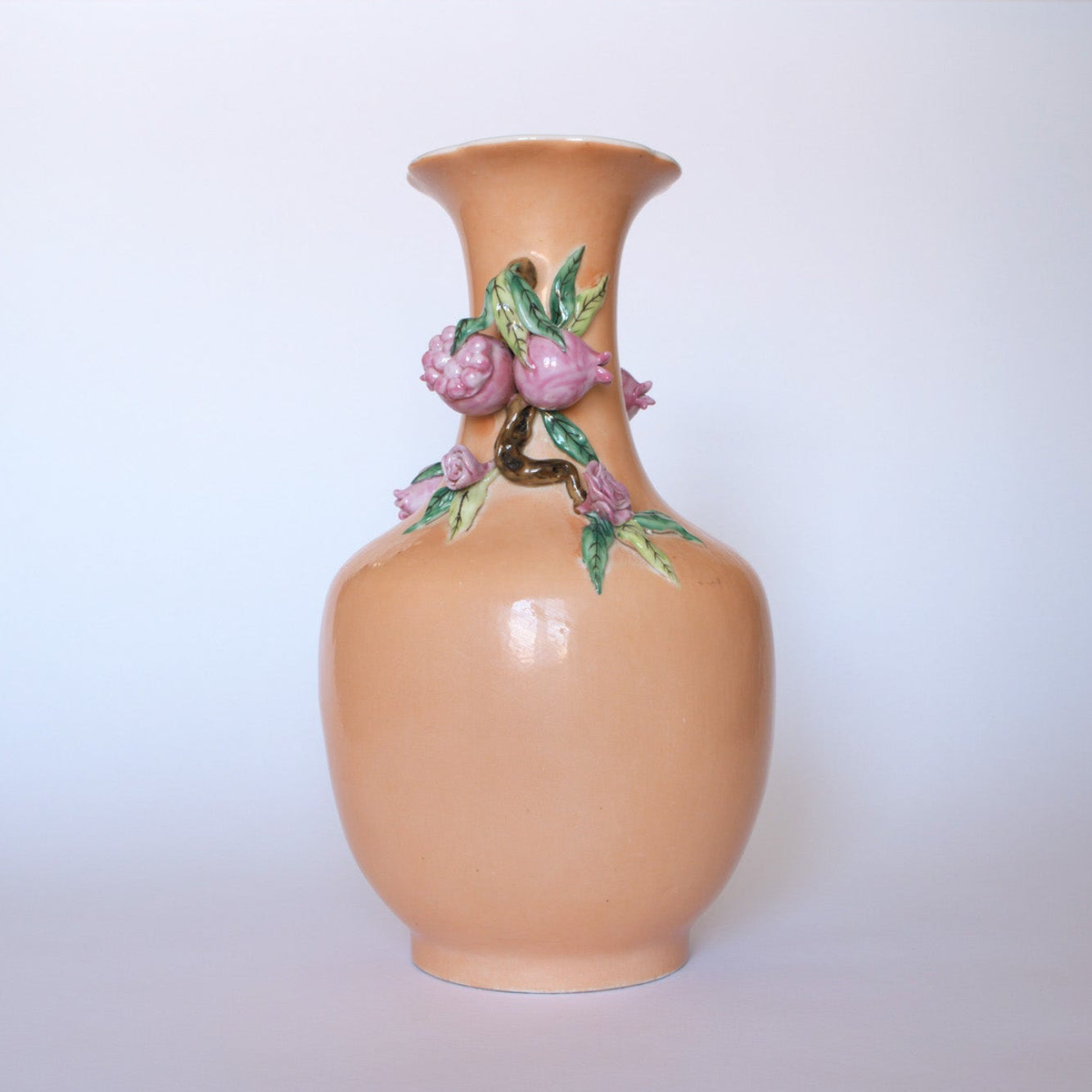 The Euro Trash Vintage Hand-Painted Floral Ceramic Vase features a peach finish, narrow neck, and raised pink flowers with green leaves wrapping around the top against a light background.