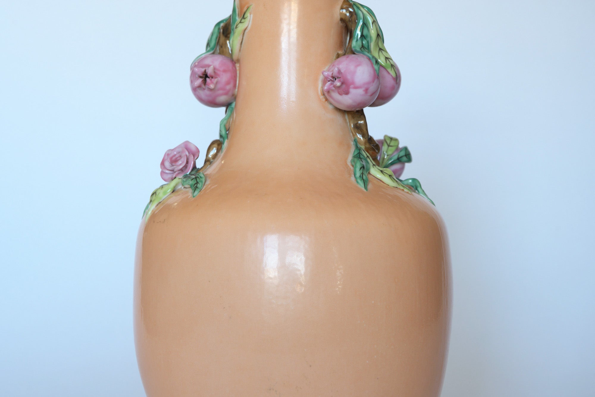 The Euro Trash Vintage Hand-Painted Floral Ceramic Vase is a beige piece with a glossy finish, adorned with pink rose-shaped details and green leaves around the neck, displayed on a light blue background.