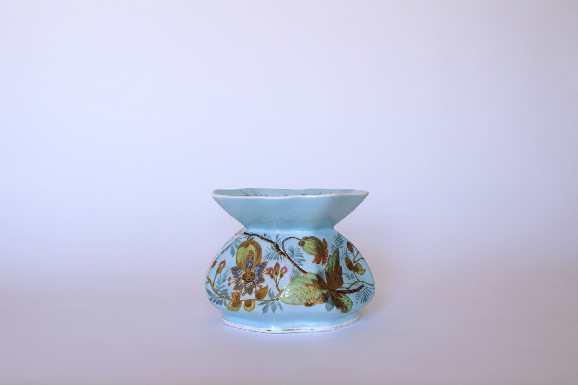 The Euro Trash Vintage Blue Floral Porcelain Vase features a light blue finish, wide flared rim, and hand-painted gold and brown floral design on a pale background.