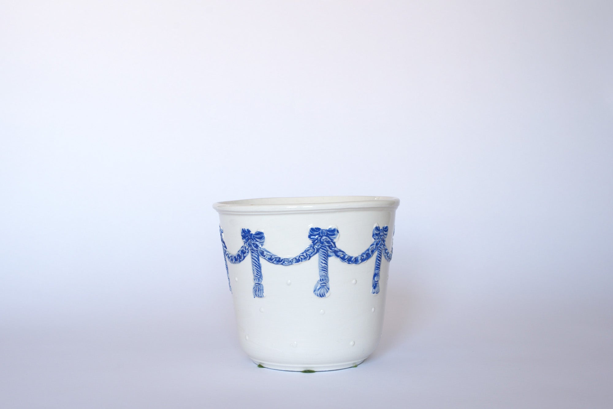 The Euro Trash Vintage White & Blue Ribbon Ceramic Cachepot features raised blue garland and bow designs, adding European-inspired charm to any plain light background.