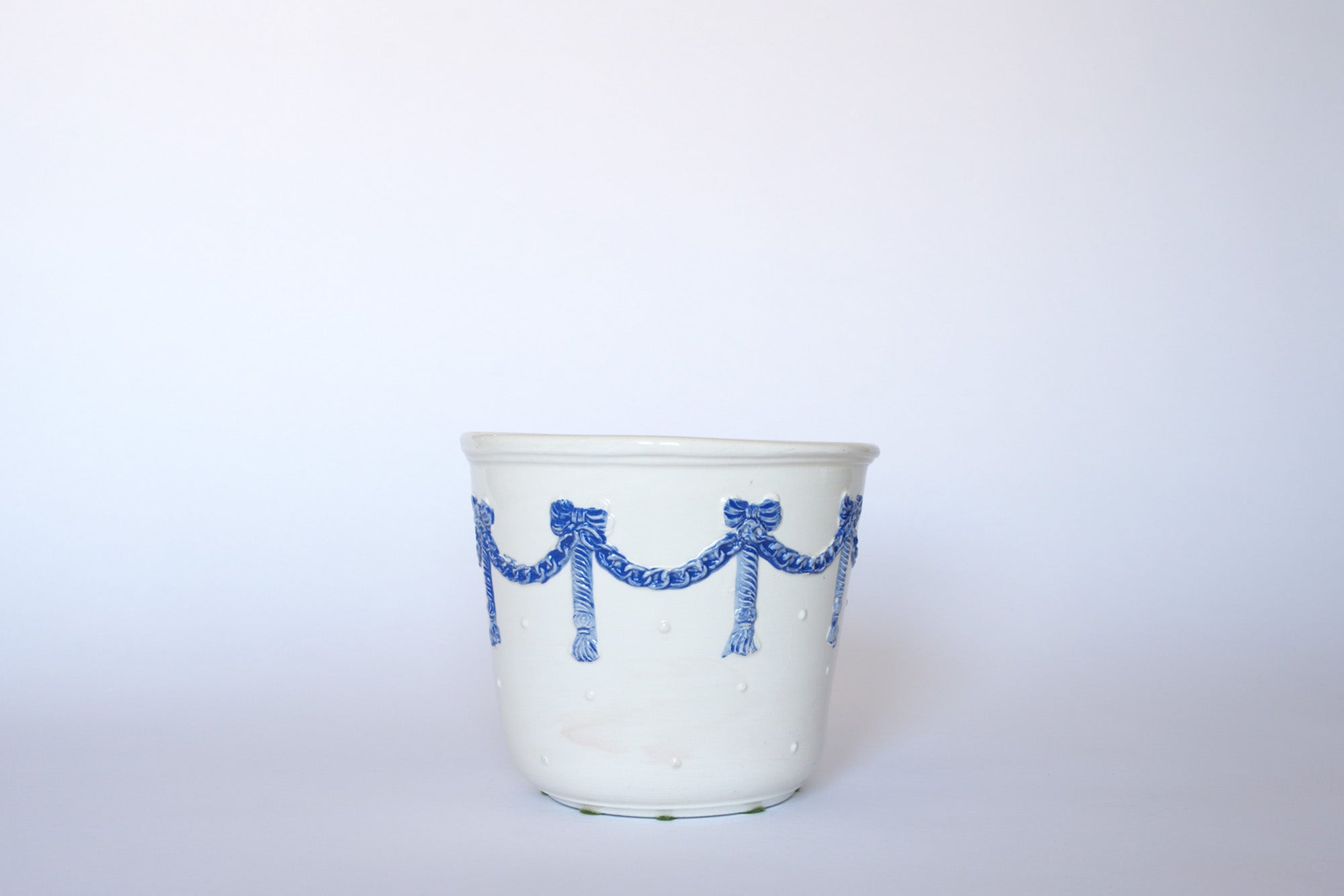 The Euro Trash Vintage White & Blue Ribbon Ceramic Cachepot features a white ceramic body with raised blue ribbon and tassel details, adding timeless charm to any plain light background.