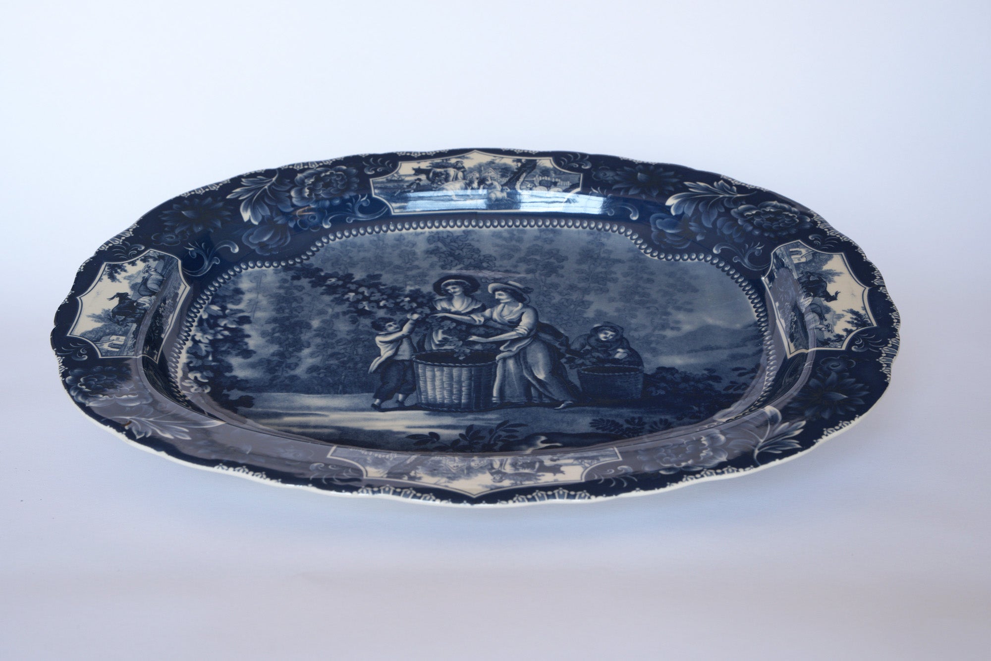 The Euro Trash Vintage Blue Transferware Oval Serving Platter features an ornate border and a central pastoral scene of three people gathering grapes—an elegant addition to any collection.