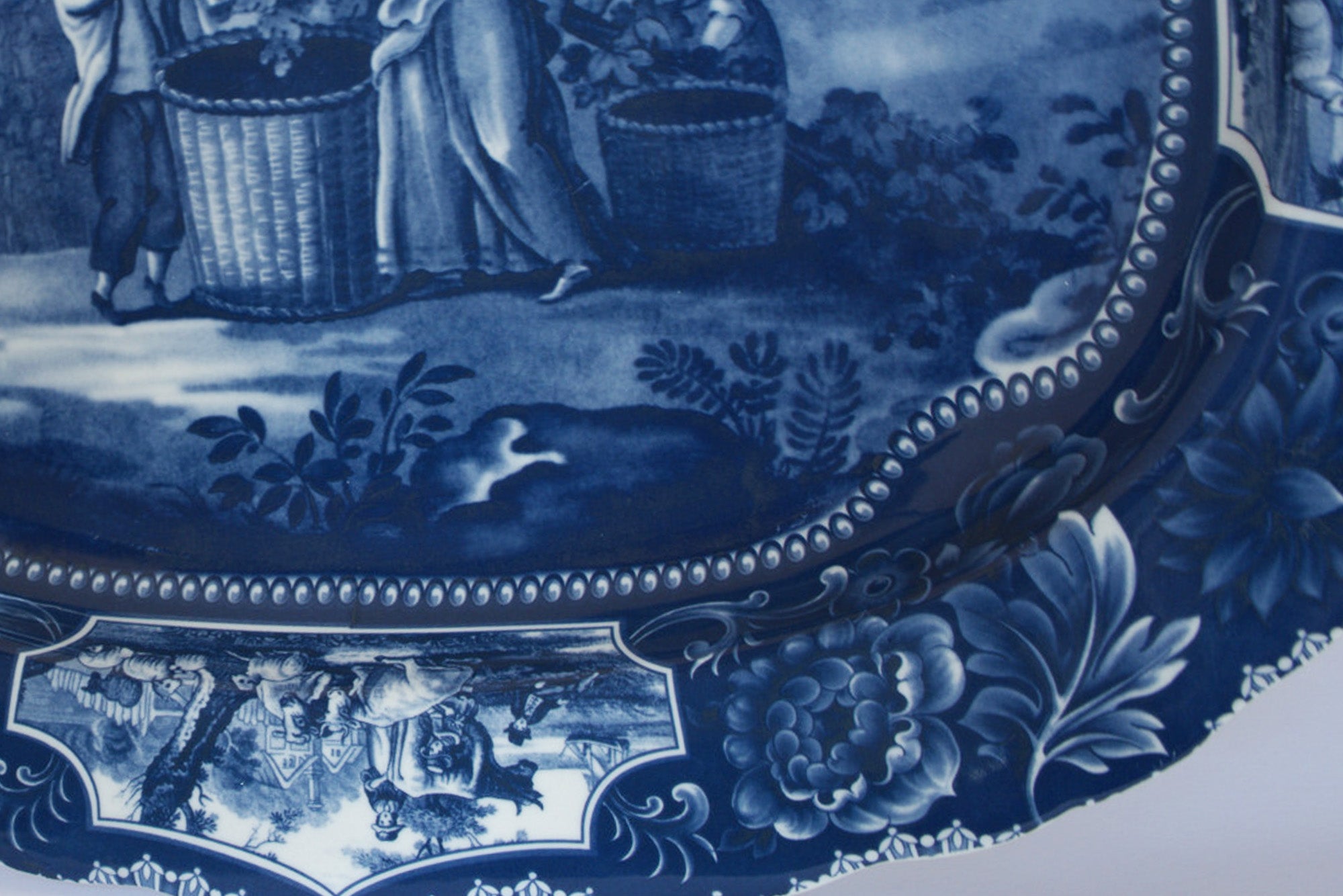 The Euro Trash Vintage Blue Transferware Oval Serving Platter features a pastoral scene with figures harvesting into baskets, intricate floral and scrollwork accents on the rim, and classic English transferware style.