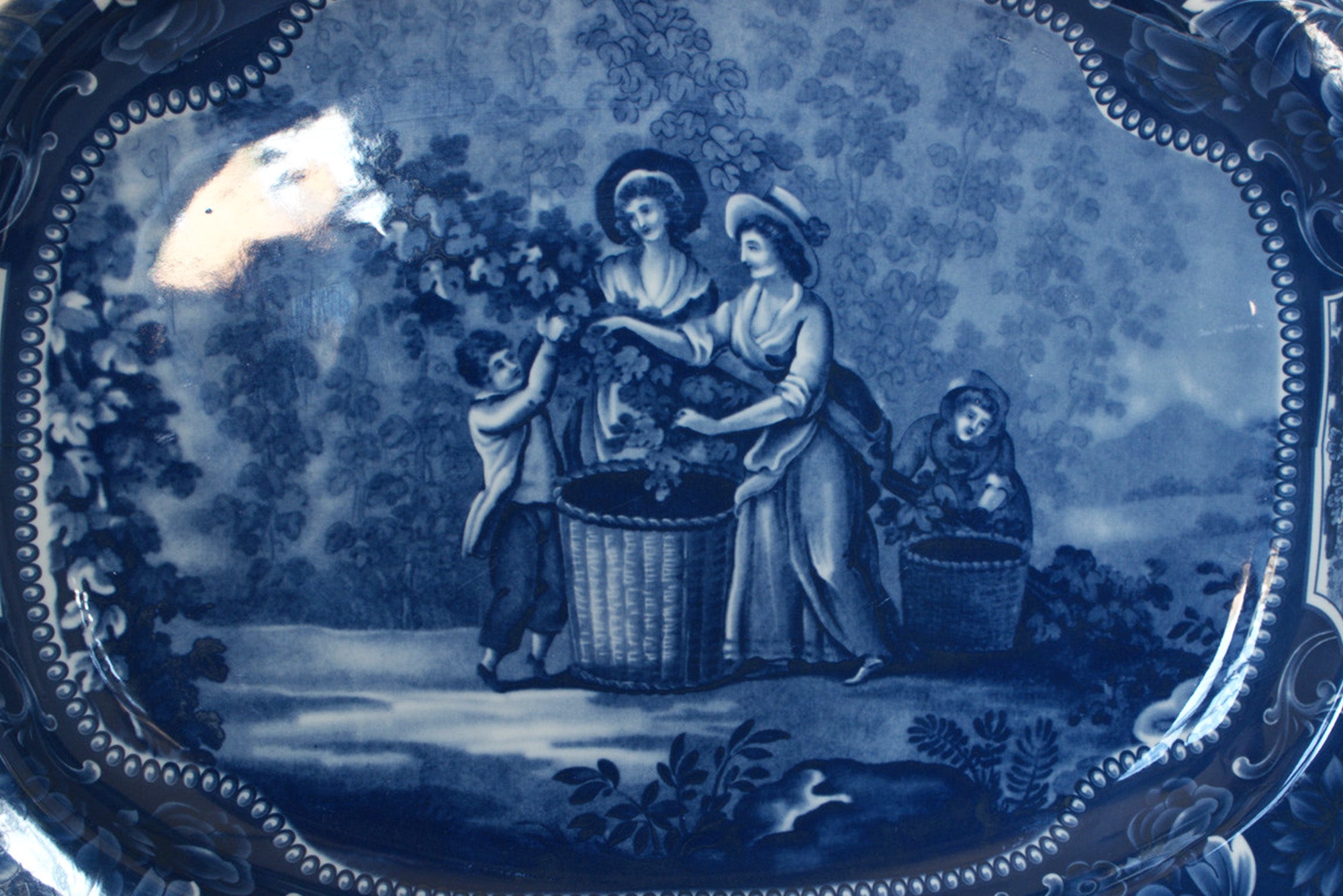 The Euro Trash Vintage Blue Transferware Oval Serving Platter features a charming pastoral scene with figures gathering grapes, ornate borders, and trees—perfect for adding classic English decor to your table.