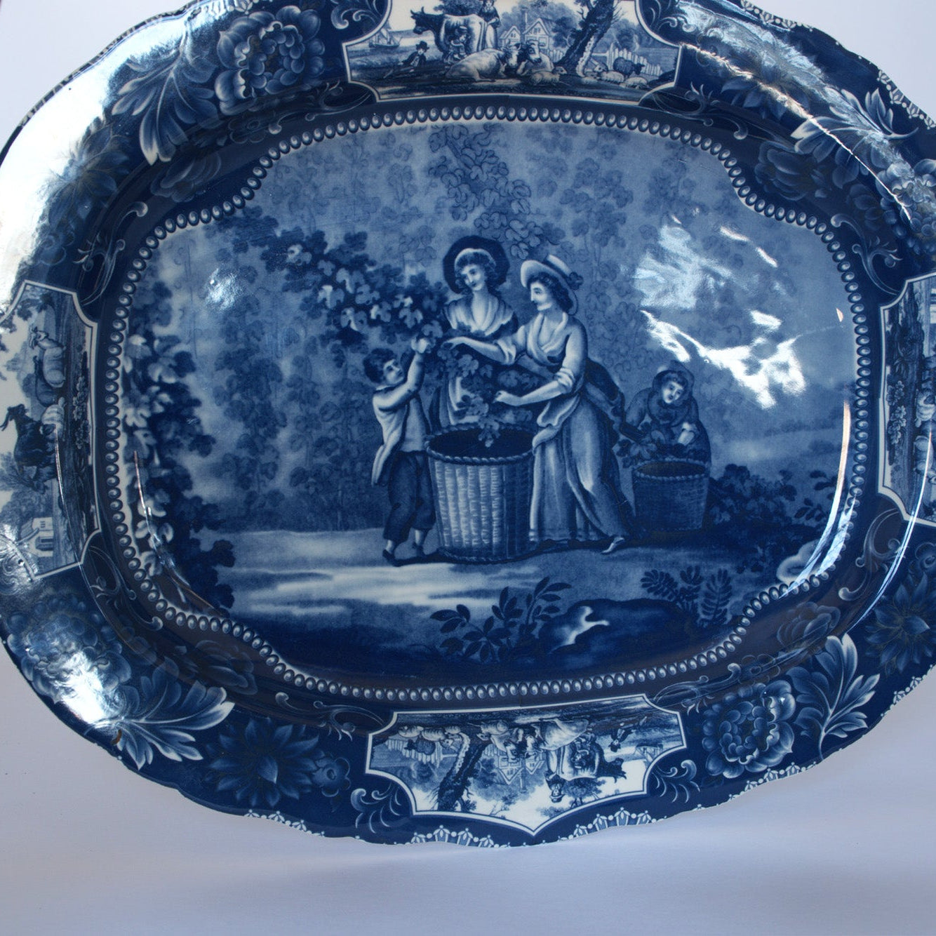 The Euro Trash Vintage Blue Transferware Oval Serving Platter features an ornate border and a pastoral scene of two women and a child harvesting grapes—a charming example of classic English transferware.