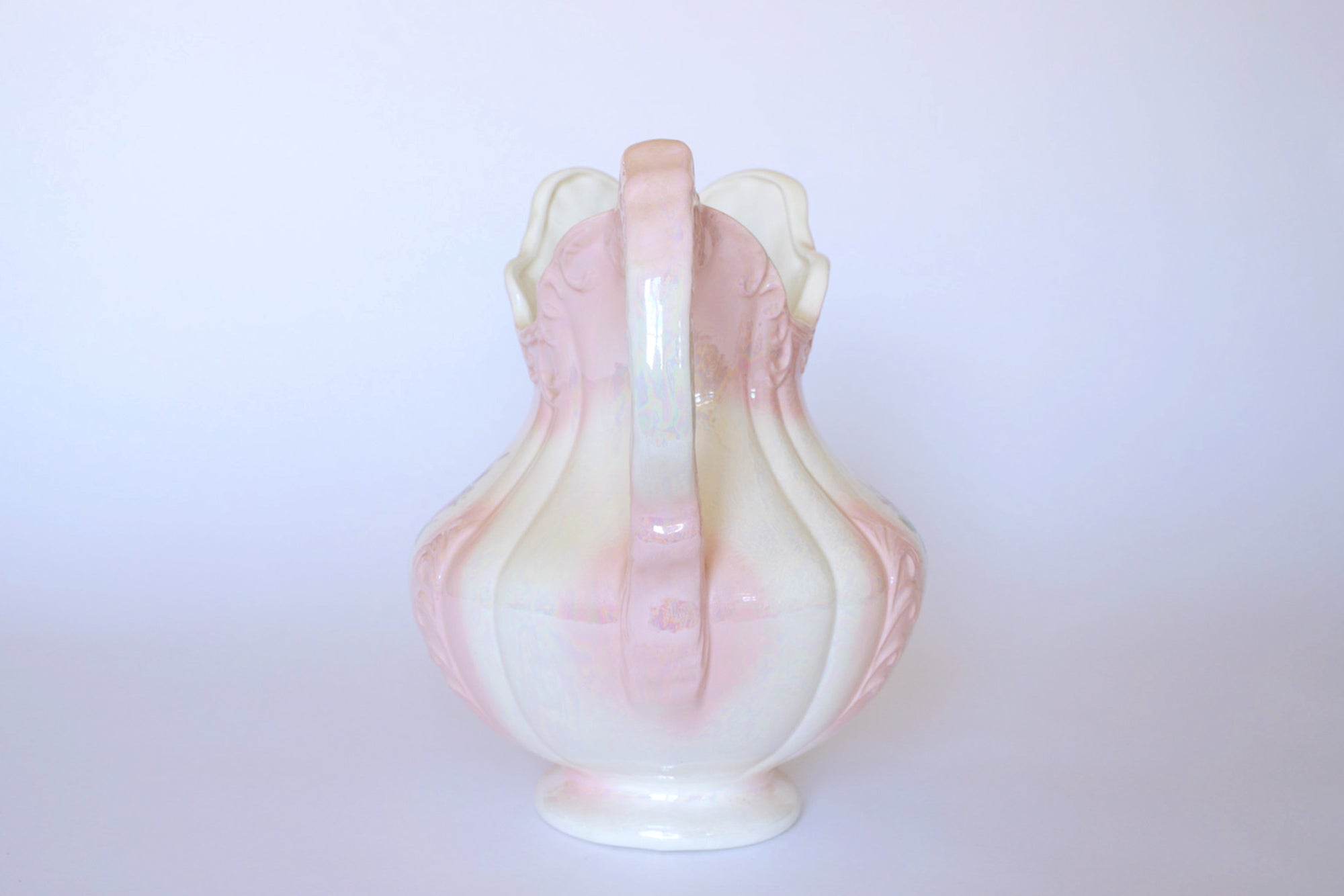 The Euro Trash Vintage Pink Lustreware Floral Ceramic Pitcher, featuring a curvy handle and pale pink and white hues with a lustre glaze, is centered on a plain white background and shown from the back.