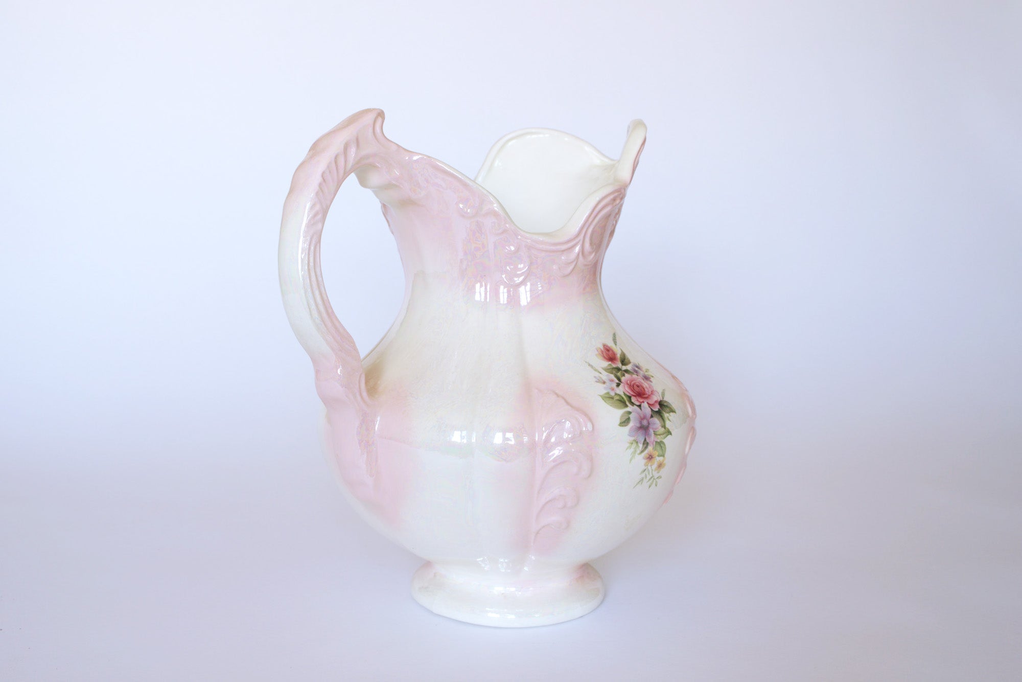 The Euro Trash Vintage Pink Lustreware Floral Ceramic Pitcher features a curved handle, hand-painted floral designs on one side, and a pastel pink lustre glaze against a plain white background.