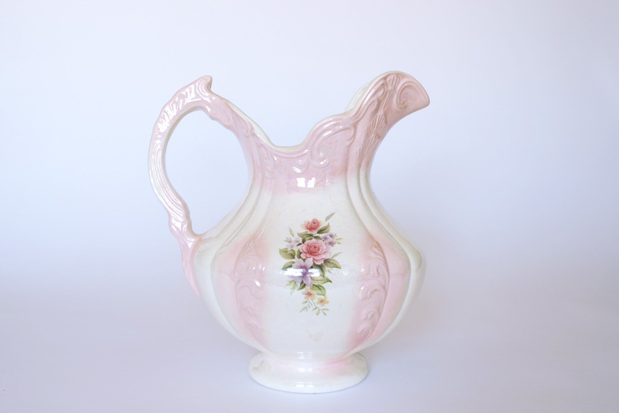 The Euro Trash Vintage Pink Lustreware Floral Ceramic Pitcher features a hand-painted pink and white floral design, ornate detailing, curved handle, and pink lustre glaze set against a plain light background.