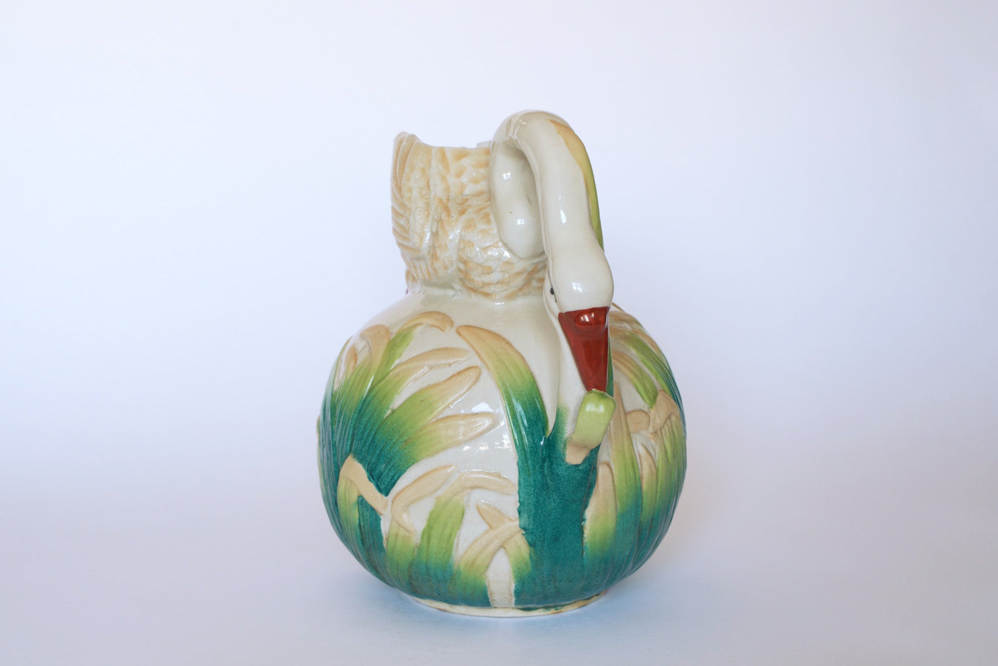 The Euro Trash Vintage Swan Motif Ceramic Pitcher features a swan-shaped handle and hand-painted green and yellow cattail designs, all set on a pure white background.