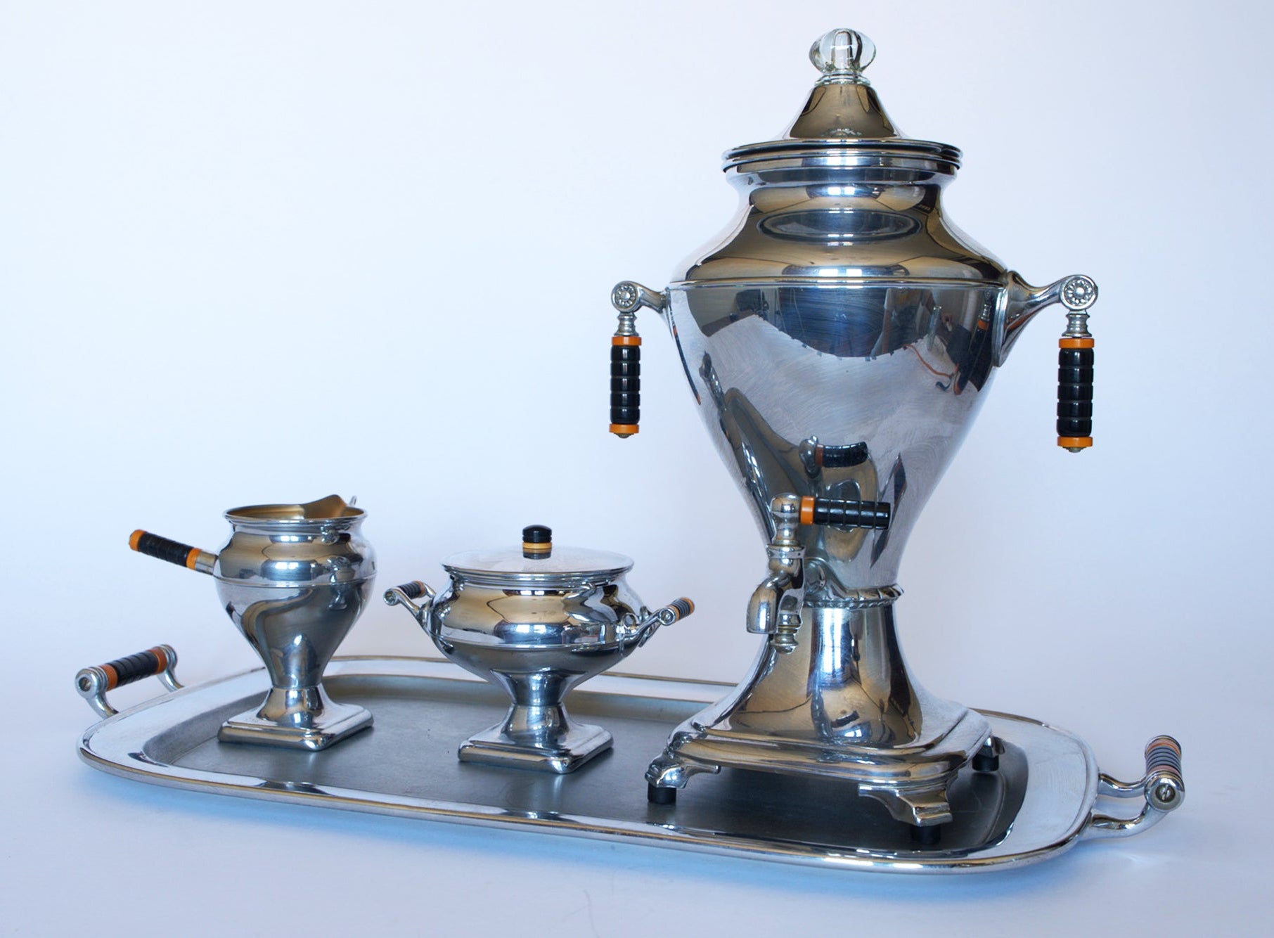 The Vintage Manning Bowman Chrome Coffee & Tea Service Set by Euro Trash features a samovar, lidded sugar bowl, and creamer with black and gold handles, displayed on a tray against a plain white background.