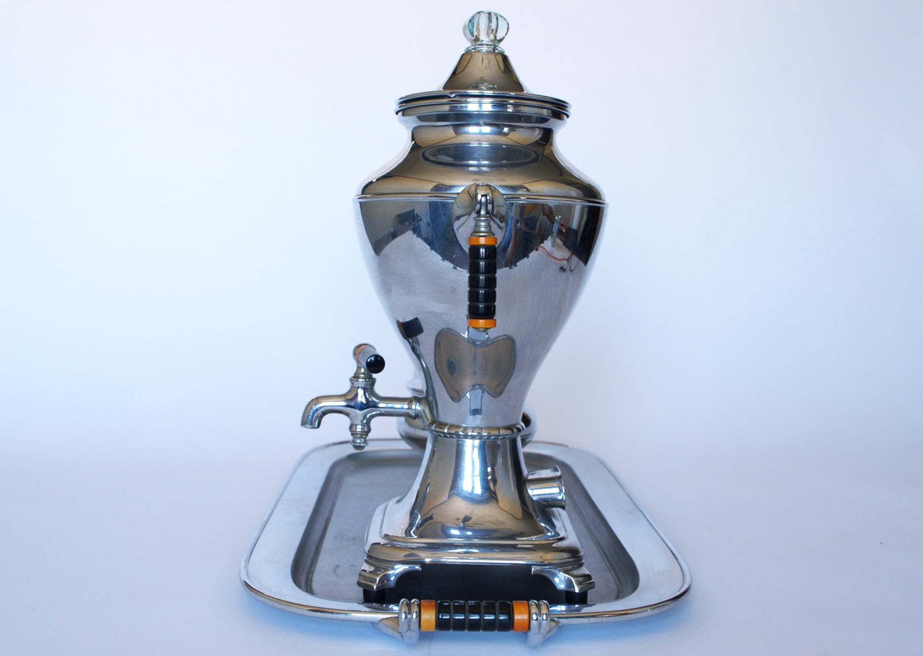 The Vintage Manning Bowman Chrome Coffee & Tea Service Set by Euro Trash features a polished, rounded samovar with black and orange accents, spout, and glass knob, displayed on a matching tray against a white background.