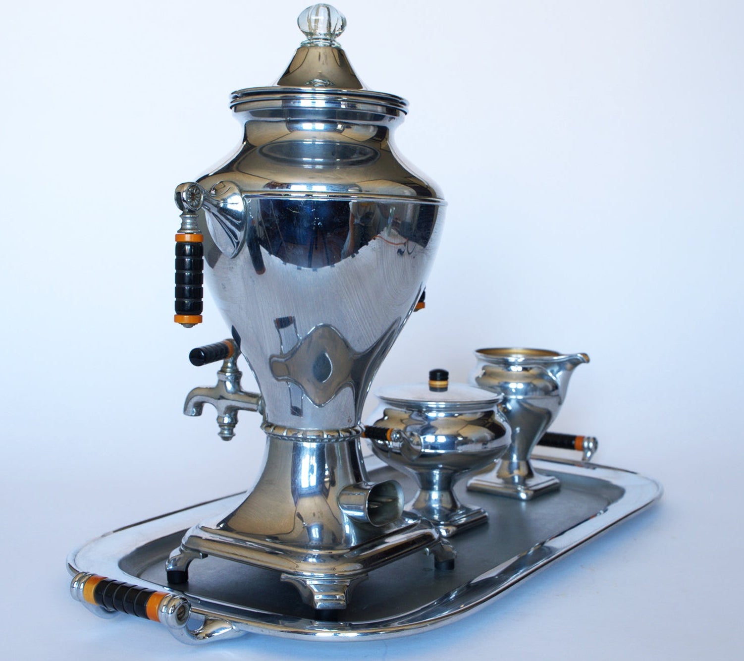 A Vintage Manning Bowman Chrome Coffee & Tea Service Set by Euro Trash, featuring black and orange handles, sits on a matching tray with sugar bowl and creamer against a plain white background.