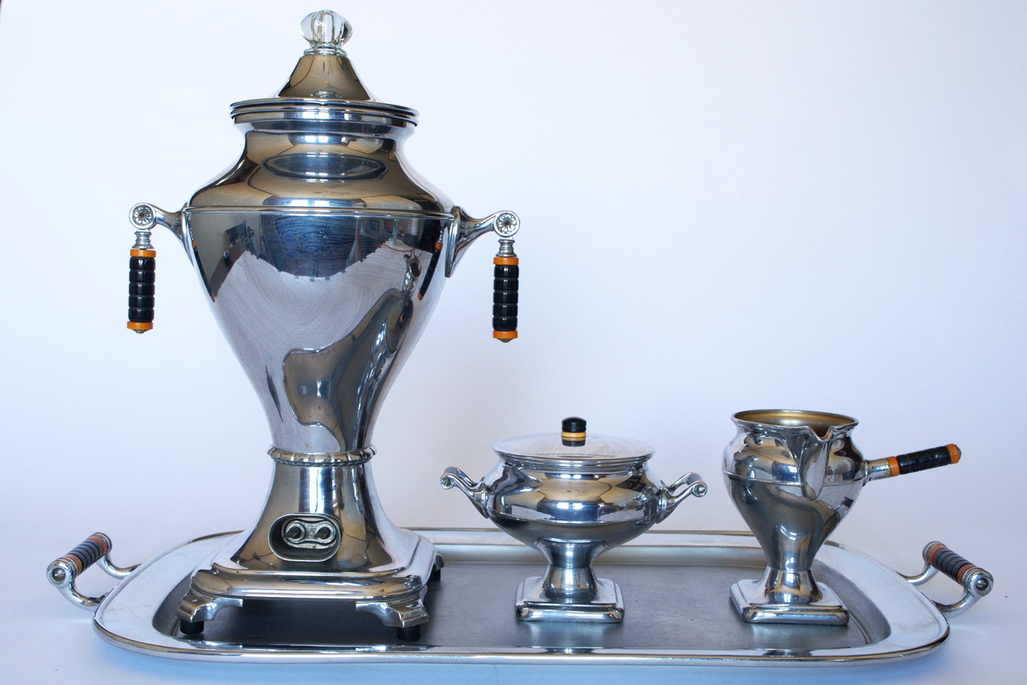 The Vintage Manning Bowman Chrome Coffee & Tea Service Set by Euro Trash features a polished Art Deco design with black and orange handles, reflective surfaces, and includes a large urn, lidded sugar bowl, creamer, and matching tray.