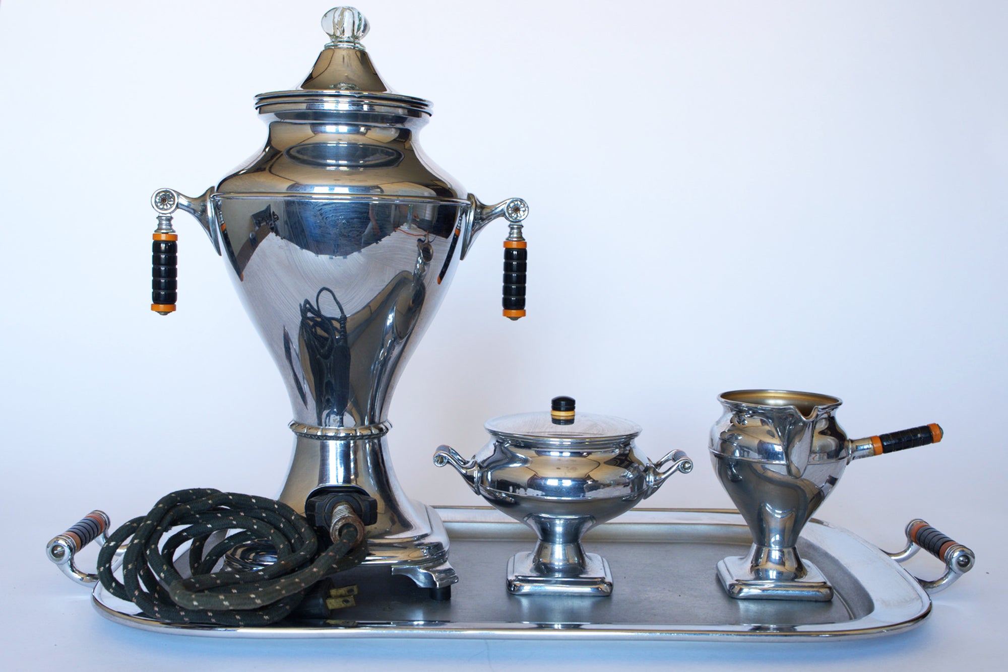 A polished Vintage Manning Bowman Chrome Coffee & Tea Service Set by Euro Trash features a samovar with black and orange handles, attached power cord, plus sugar bowl and creamer, all displayed on a metal tray.