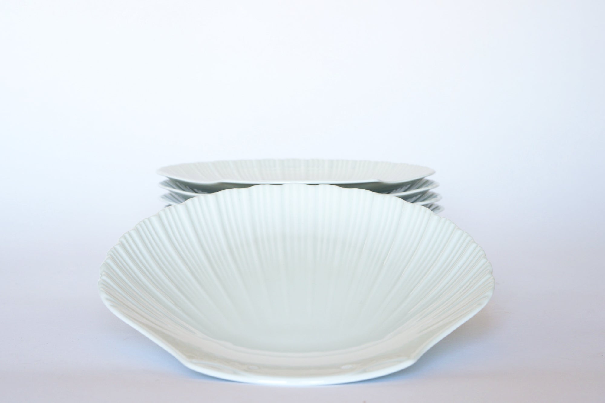 The Euro Trash Vintage White Ceramic Shell Plate Set features four shell-patterned white plates stacked in the background, with one matching plate in front on a plain white surface—ideal for coastal-inspired tableware.