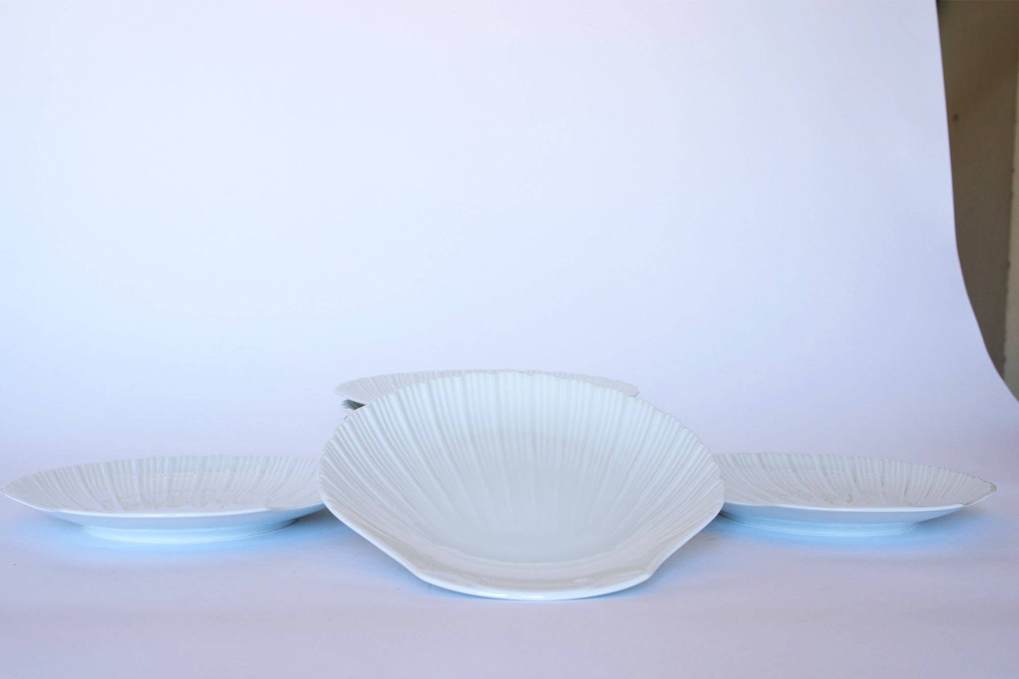 Four Vintage White Ceramic Shell Plates from Euro Trash—three small in back, one large in front—are arranged on a white surface and background, creating elegant coastal tableware for your dining setting.