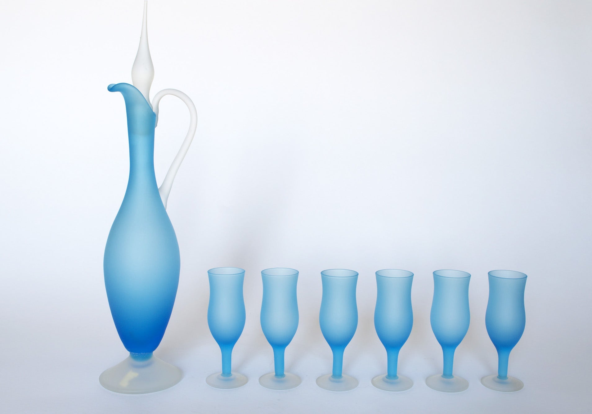 The Euro Trash Vintage Frosted Blue Glass Decanter and Stemware Set features a tall blue decanter with white handle and stopper, paired with five matching blue stemmed glasses, all displayed in a row on a white background.