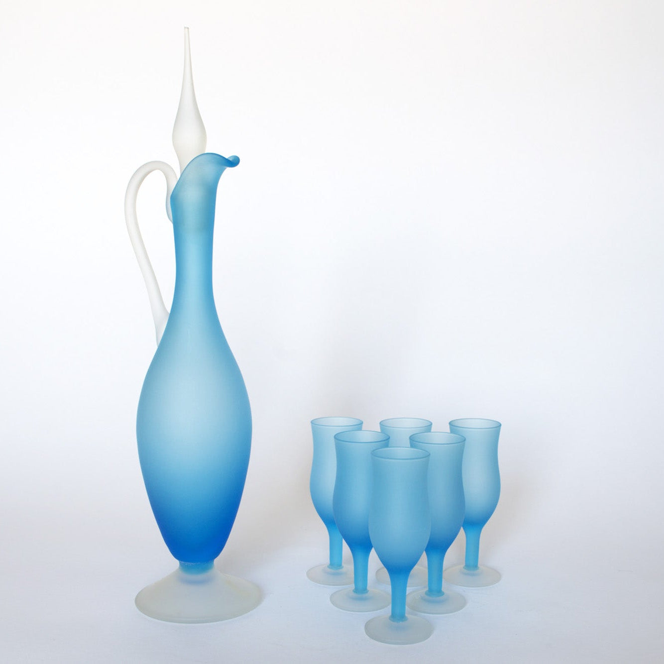 The Euro Trash Vintage Frosted Blue Glass Decanter and Stemware Set features a tall decanter with handle and stopper beside six elegant blue stemmed glasses, displayed on a plain white background.