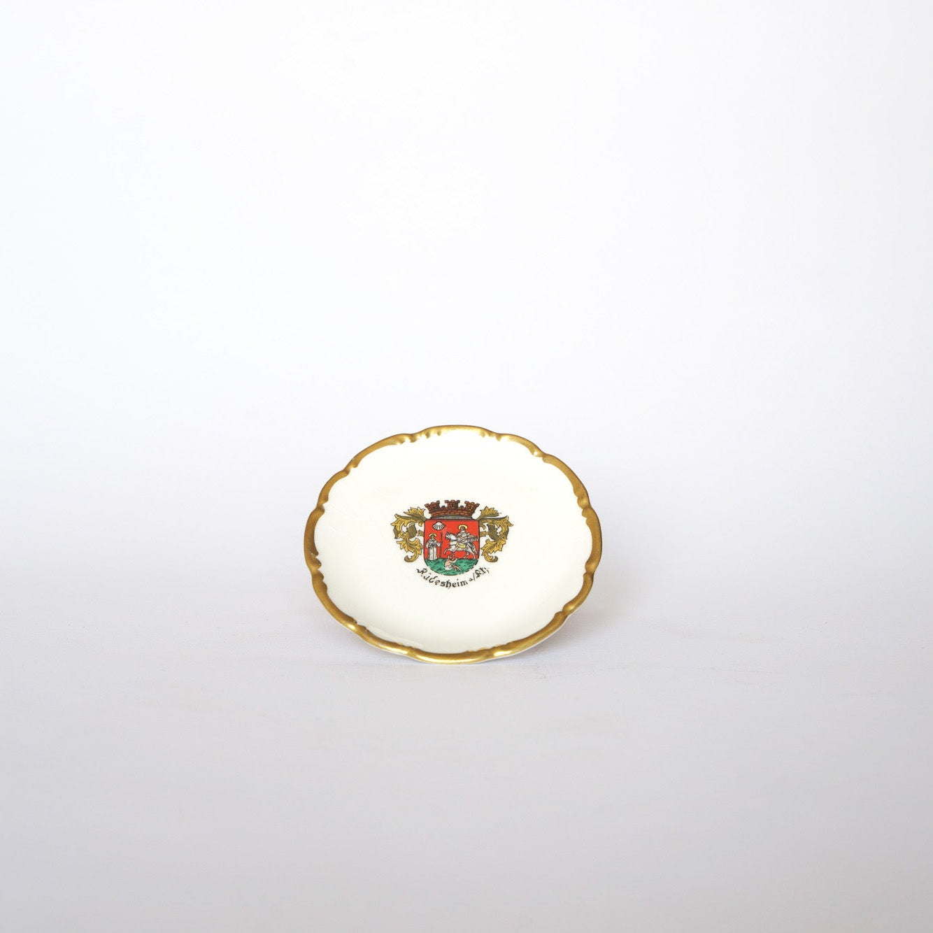 Euro Trash Vintage Crest Porcelain Trinket Dish features a gold rim and a vibrant heraldic crest with a red shield and ornate details at the center, set on a plain white background.