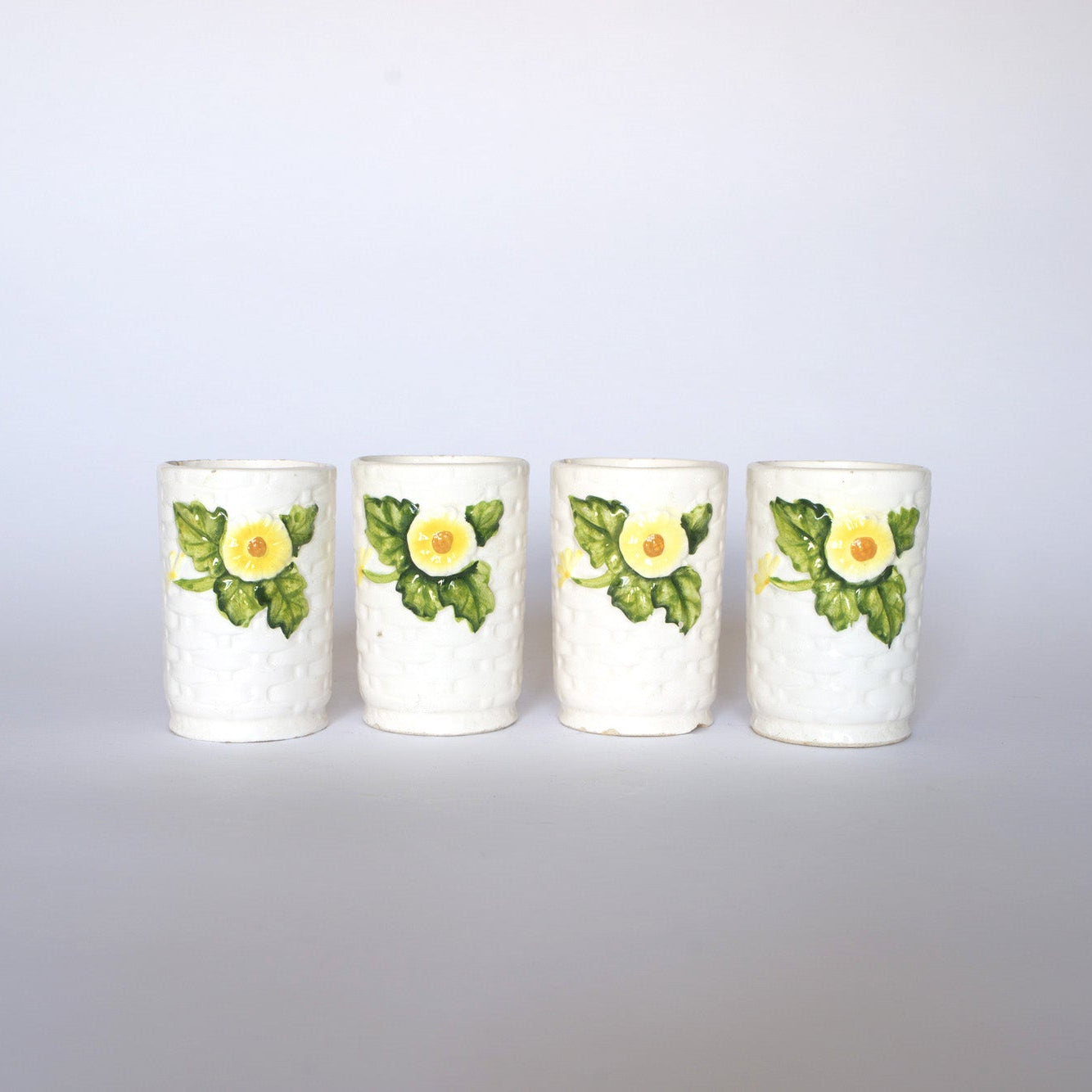 The Euro Trash Vintage Ivy Motif Ceramic Bud Vases feature four white ceramic cups with textured surfaces, each adorned with a yellow flower and green leaves, evoking vintage charm and displayed in a row against a white background.