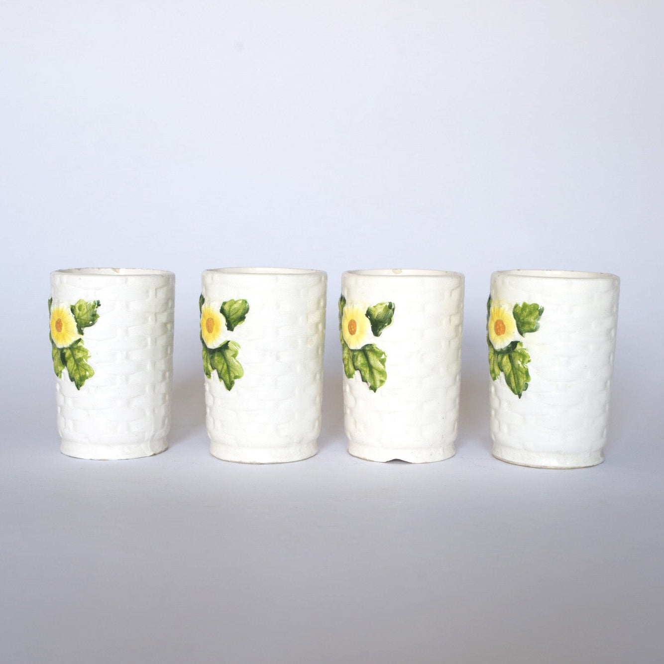 Euro Trash’s Vintage Ivy Motif Ceramic Bud Vases—a set of four white ceramic vases with basket-weave texture, yellow flowers, and green leaves—are arranged in a row on a plain white background.