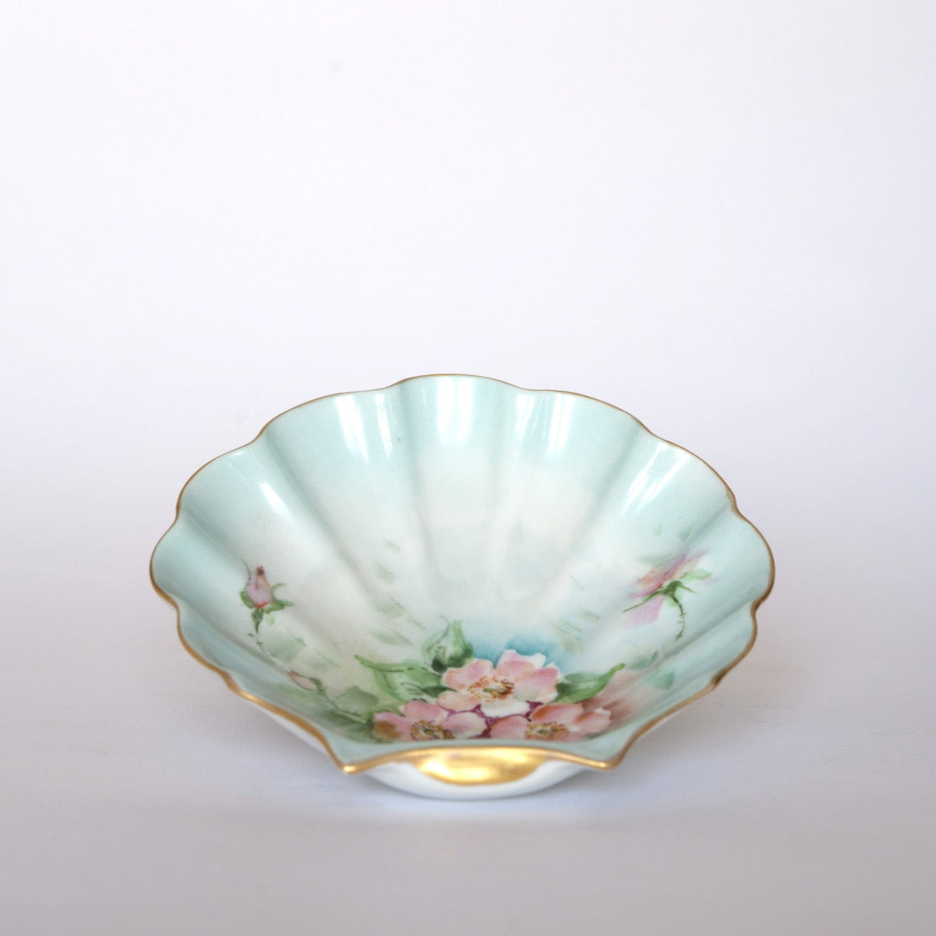 The Euro Trash Vintage Hand-Painted Floral Porcelain Plate features a scallop shape, gold rim, pink flowers, green leaves, and a pale blue-green glaze on a plain white background.