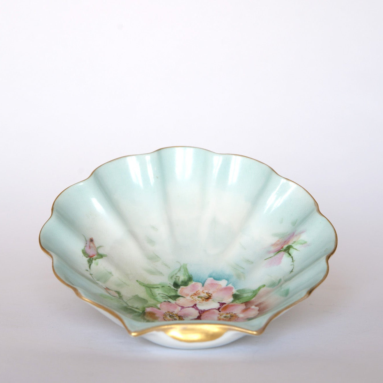 The Euro Trash Vintage Hand-Painted Floral Porcelain Plate features a scallop shape, pale blue-green glaze, gold rim, and pink flowers with green leaves, displayed on a plain white background.