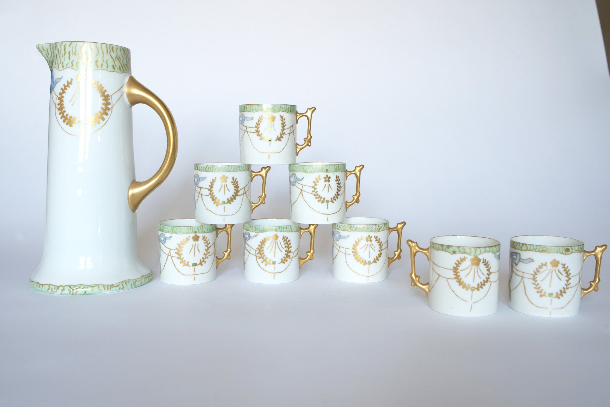 The Euro Trash Vintage White Porcelain Teacup Set with Gold Handles features a tall pitcher and eight matching cups adorned with gold and green accents, elegantly arranged in a graceful pyramid.