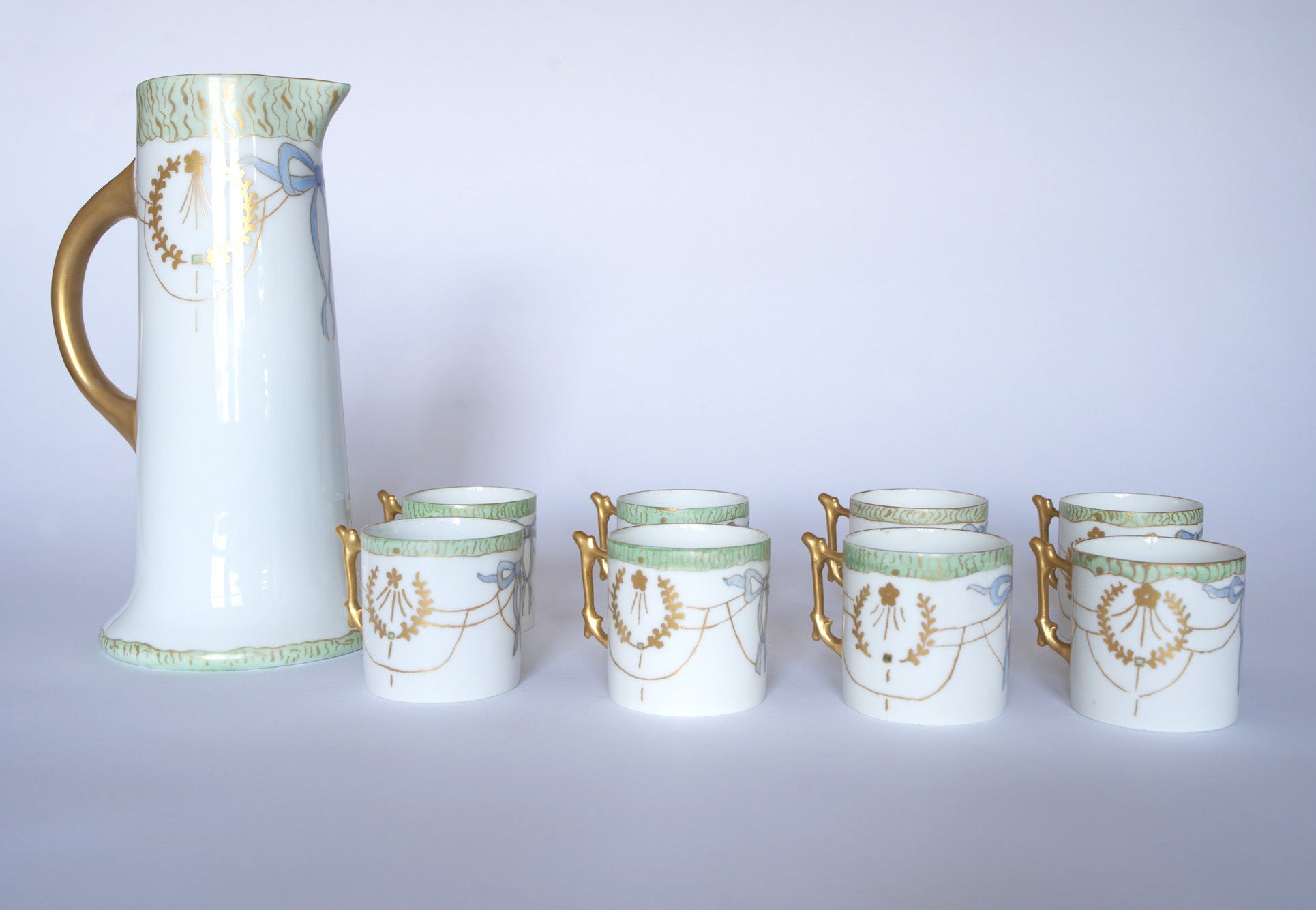 The Euro Trash Vintage White Porcelain Teacup Set with Gold Handle includes a classic white pitcher and six matching cups, each featuring green rims, gold garlands, and blue bows on a plain light background.