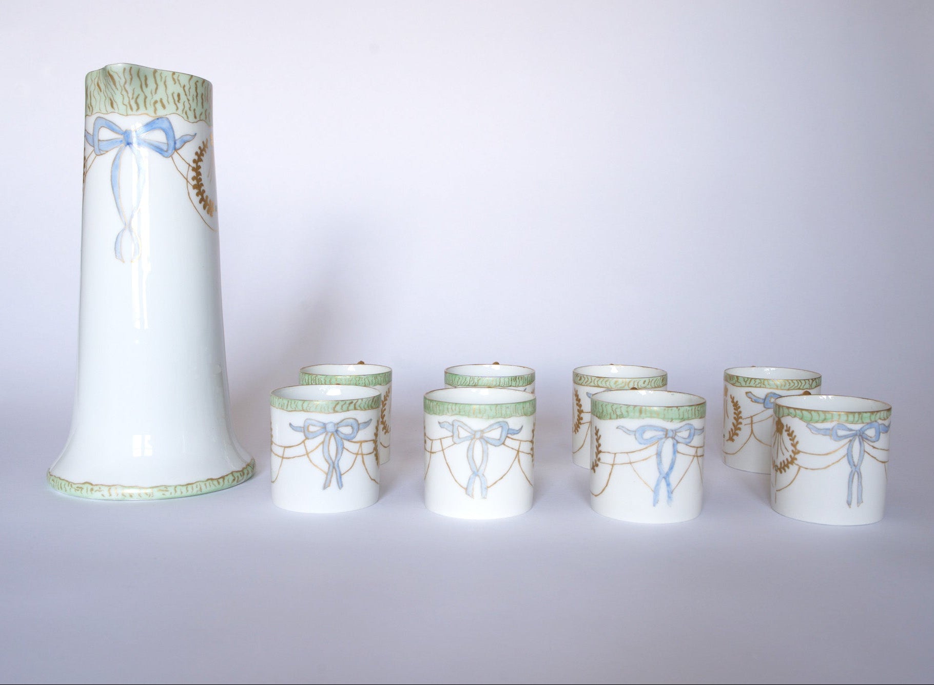 The Euro Trash Vintage White Porcelain Teacup Set with Gold Handle features white cups and a tall jug with blue ribbon, gold wheat motifs, green rims, and elegant gold handles, shown on a plain white background.