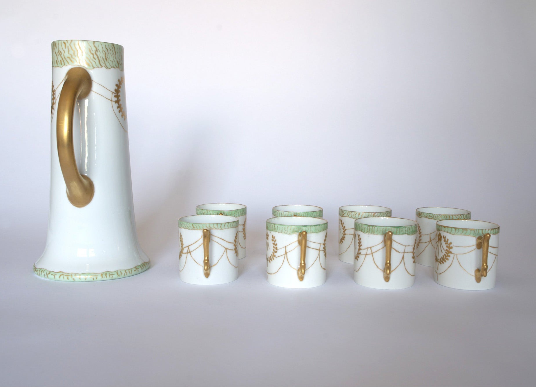 The Euro Trash Vintage White Porcelain Teacup Set with Gold Handle features a tall pitcher and eight matching teacups, each adorned with gold and green accents, all arranged on a plain white background.