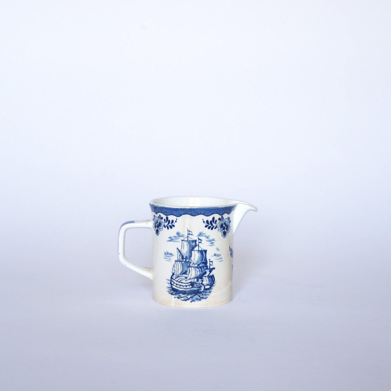 The Euro Trash Vintage Blue & White Ship Motif Porcelain Creamer features a handle, spout, elegant floral patterns, and a nautical ship design set against a crisp white background.
