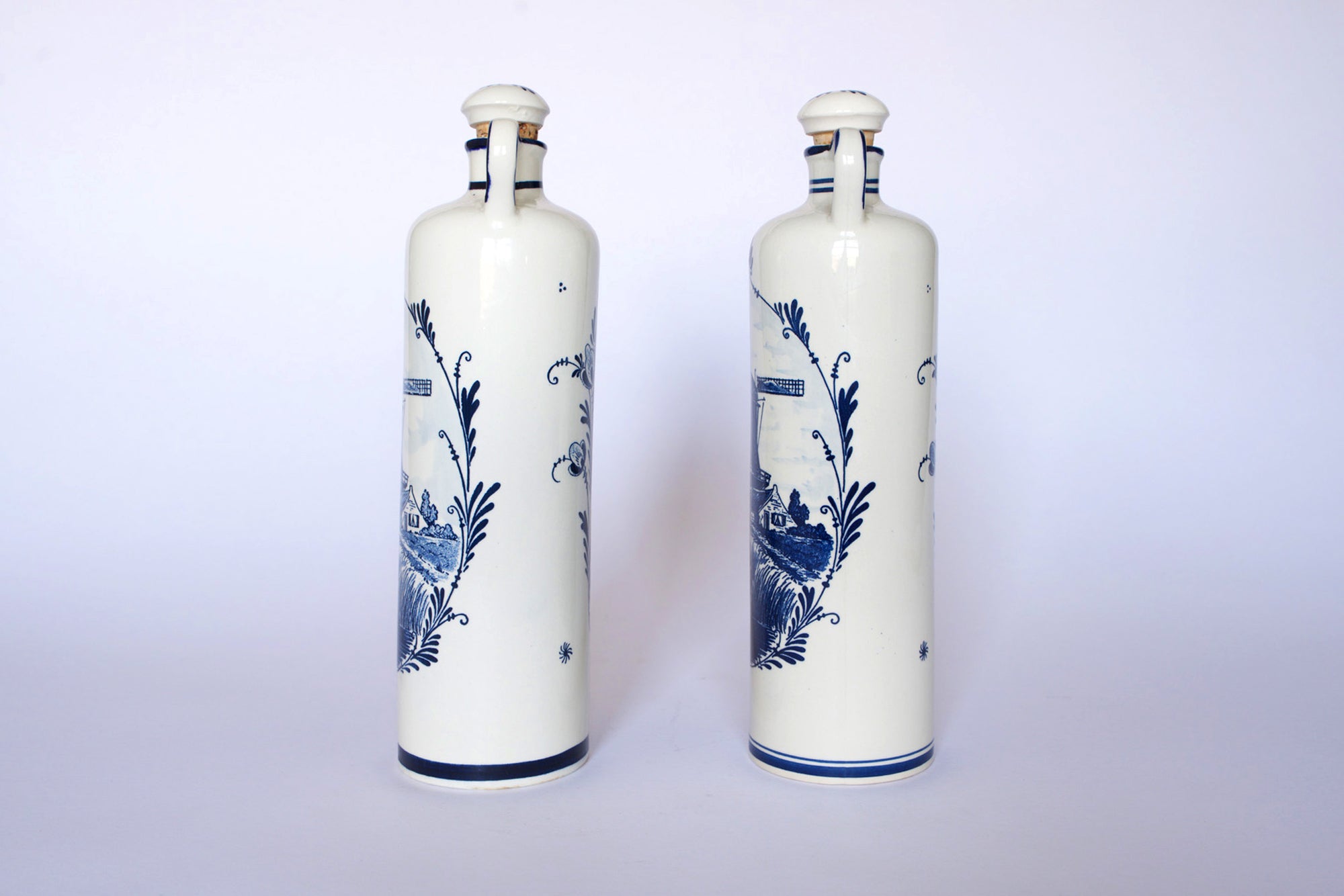 The Euro Trash Vintage Blue & White Windmill Motif Ceramic Bottle Decanters feature tall ceramic bottles with scenic windmill designs, displayed upright on a plain white background as a charming decorative set.