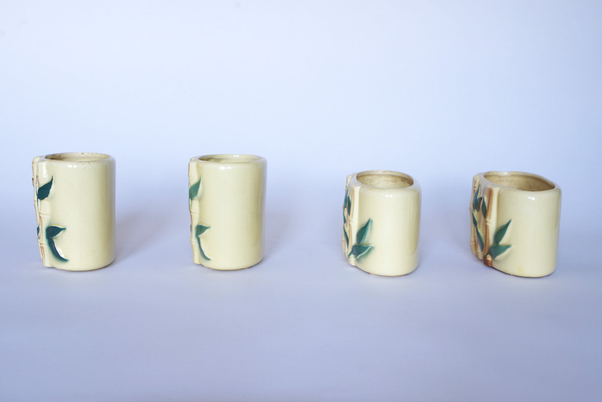 Set of four small, cylindrical Vintage Bamboo Ceramic Vases by Euro Trash, featuring pale yellow glaze with green leaf and bamboo motifs. Shown from various angles on a white background—ideal for nature-inspired decor.